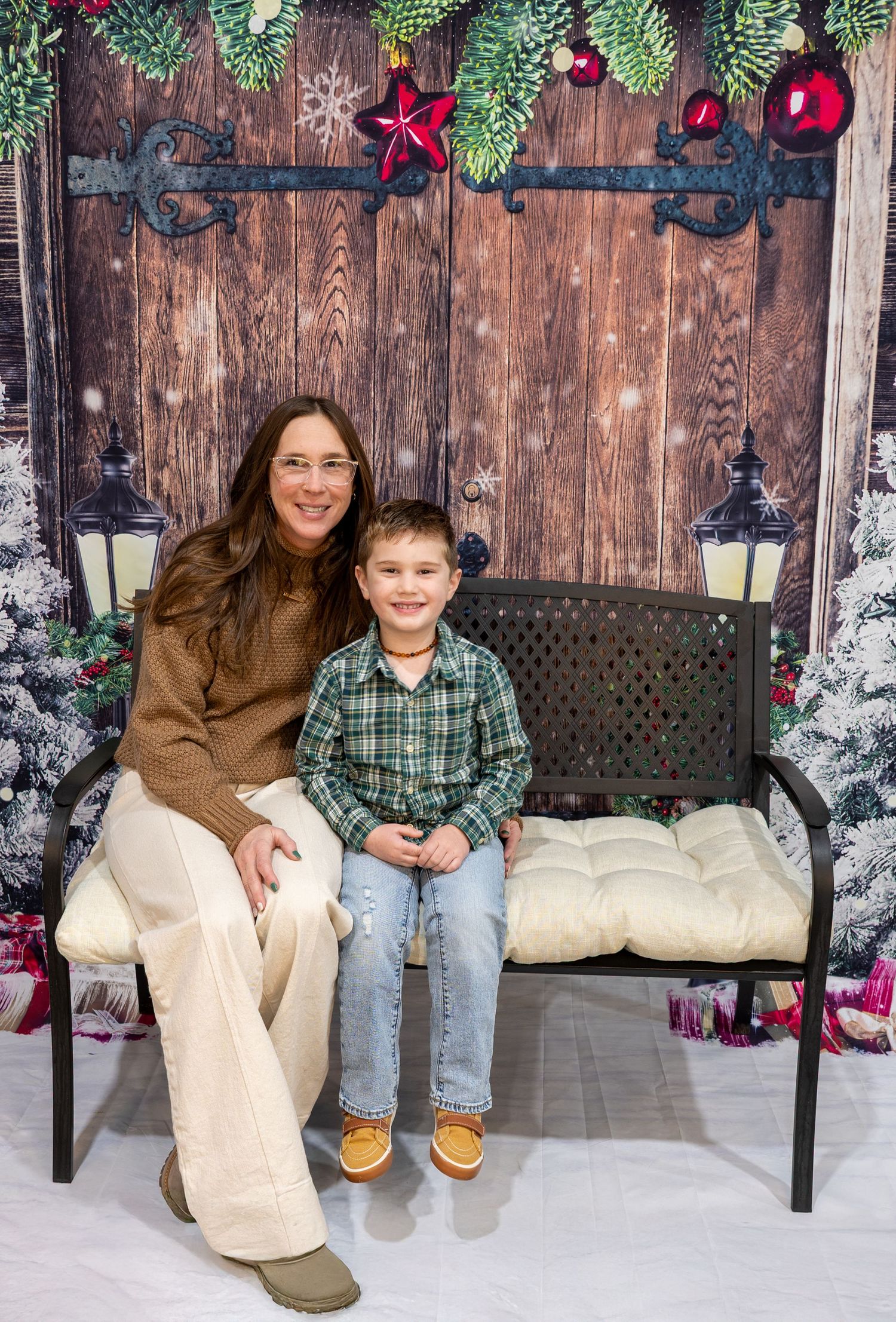 Holiday photo setup with wooden backdrop, evergreen garland, red ornaments, and bench decorated with white cushions.