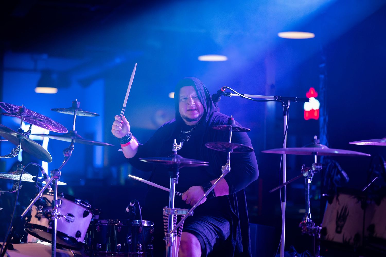 Concert stage lit with blue lights showing musicians performing with drums and instruments in a dark venue setting.