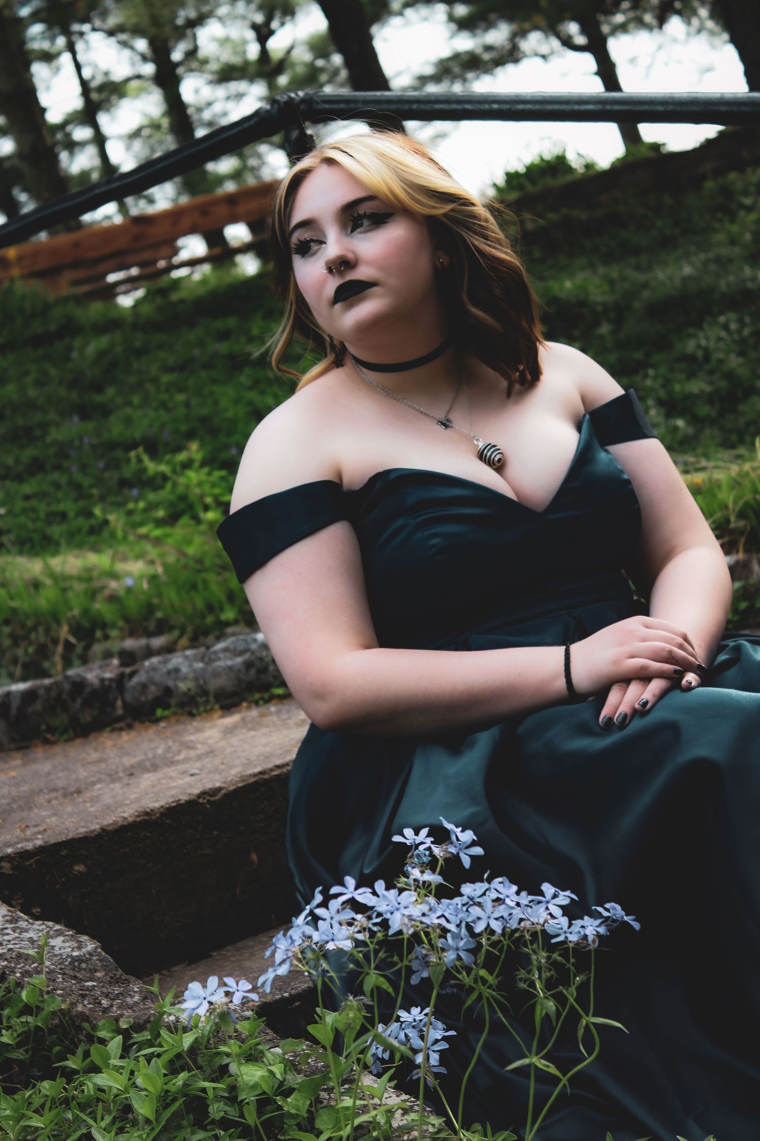 A person in a black dress lies on stone steps near delicate blue flowers in a moody outdoor setting.