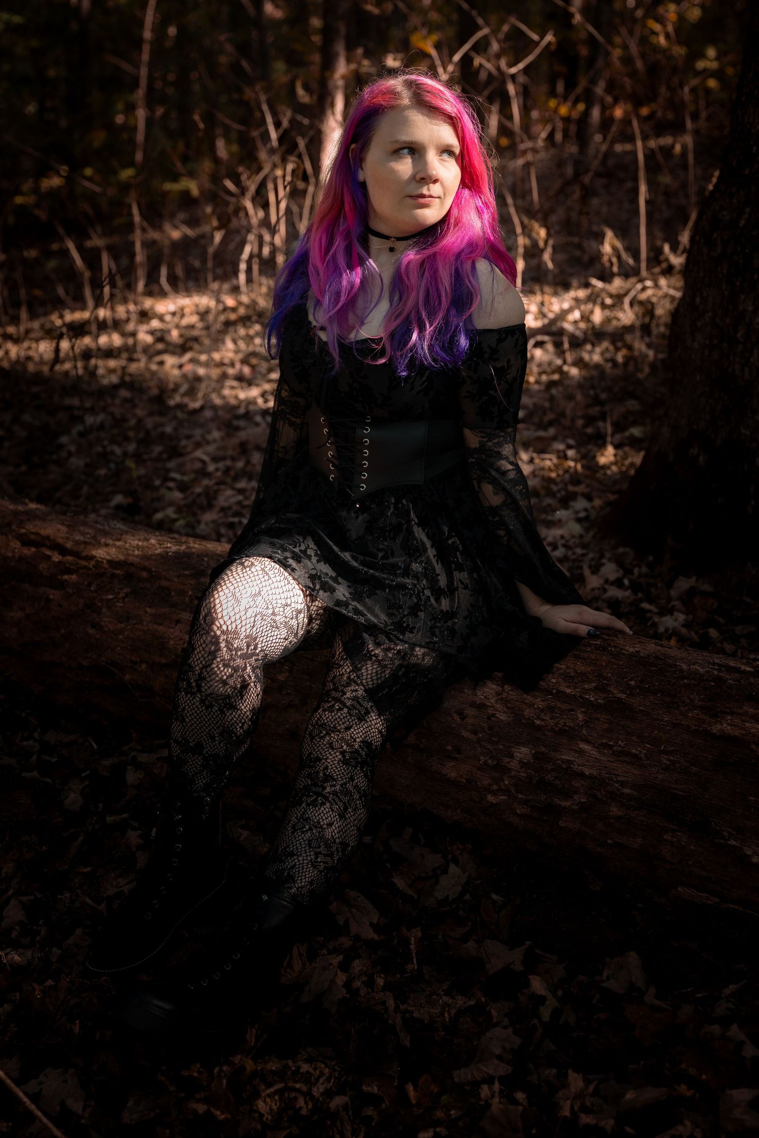 A person with vibrant pink and purple hair wearing black clothing poses in a dark forest setting.