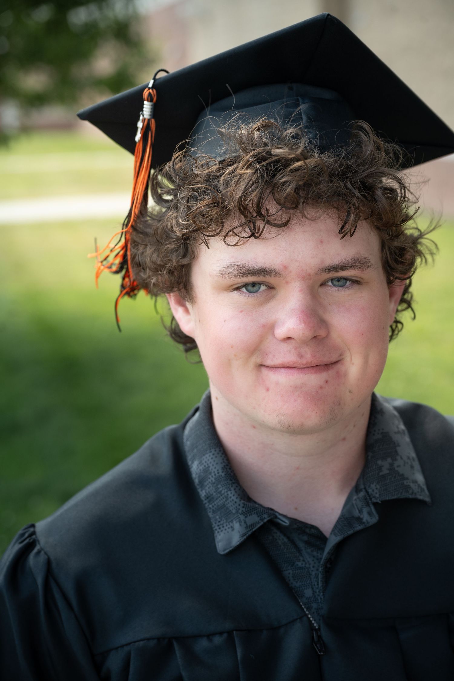 Young man in black graduation cap and gown with orange tassel smiling outdoors on graduation day.