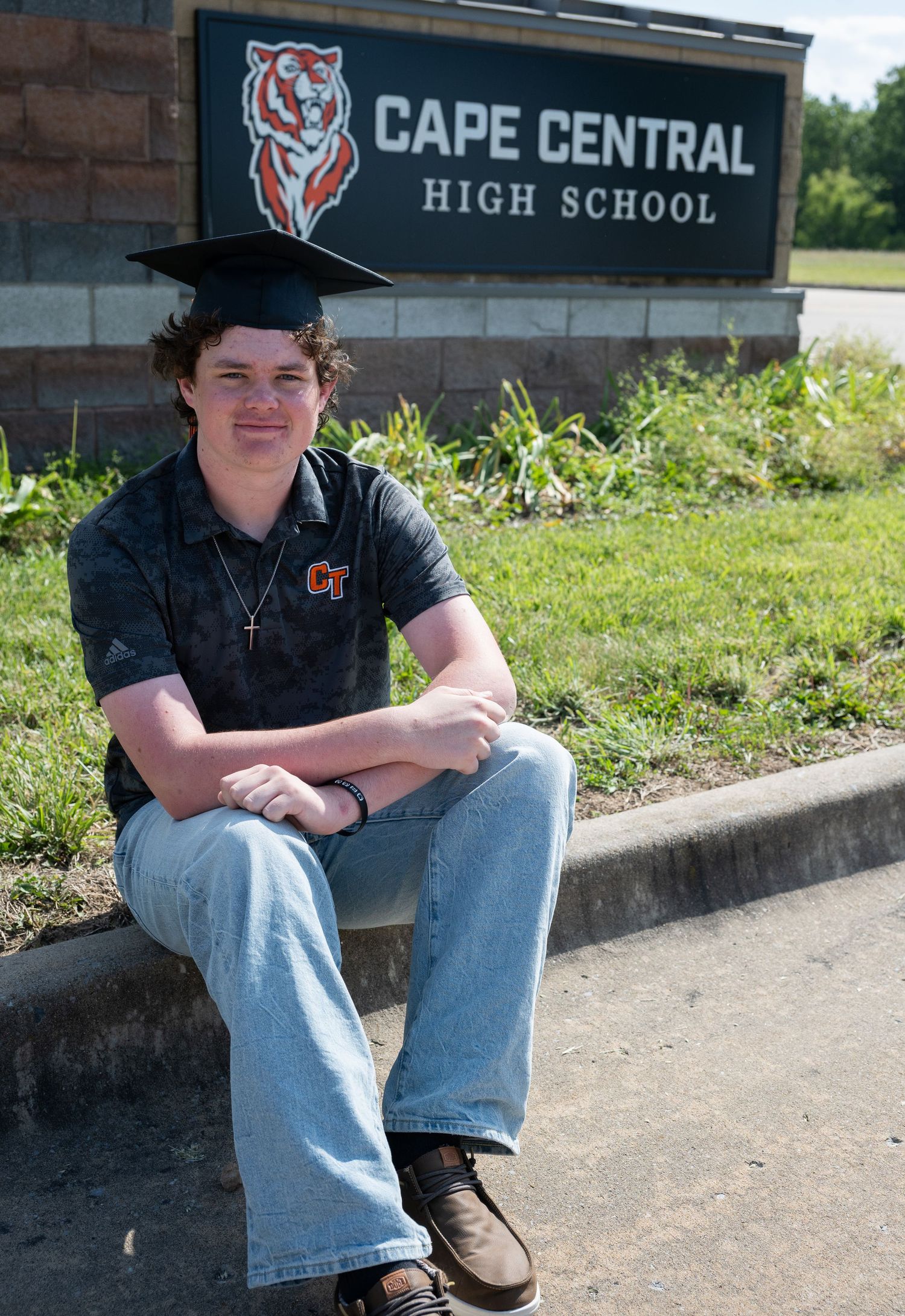 A graduating student in cap sits in front of Cape Central High School sign, wearing a school polo shirt.