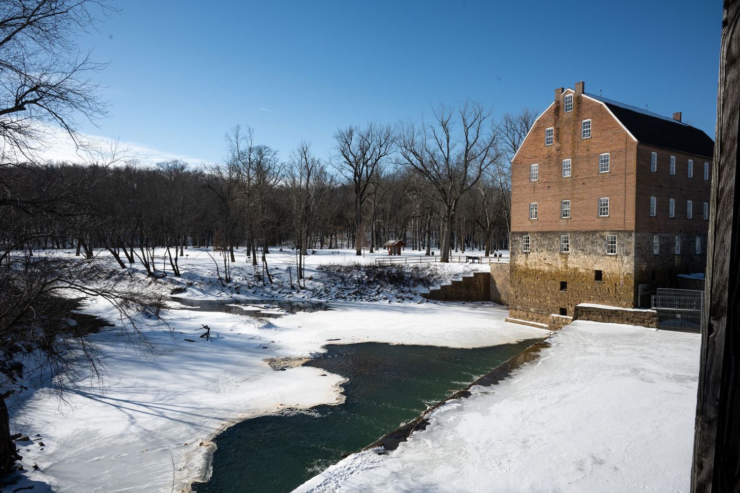 Historic stone buildings stand along a snowy riverbank in winter with bare trees dotting the landscape.