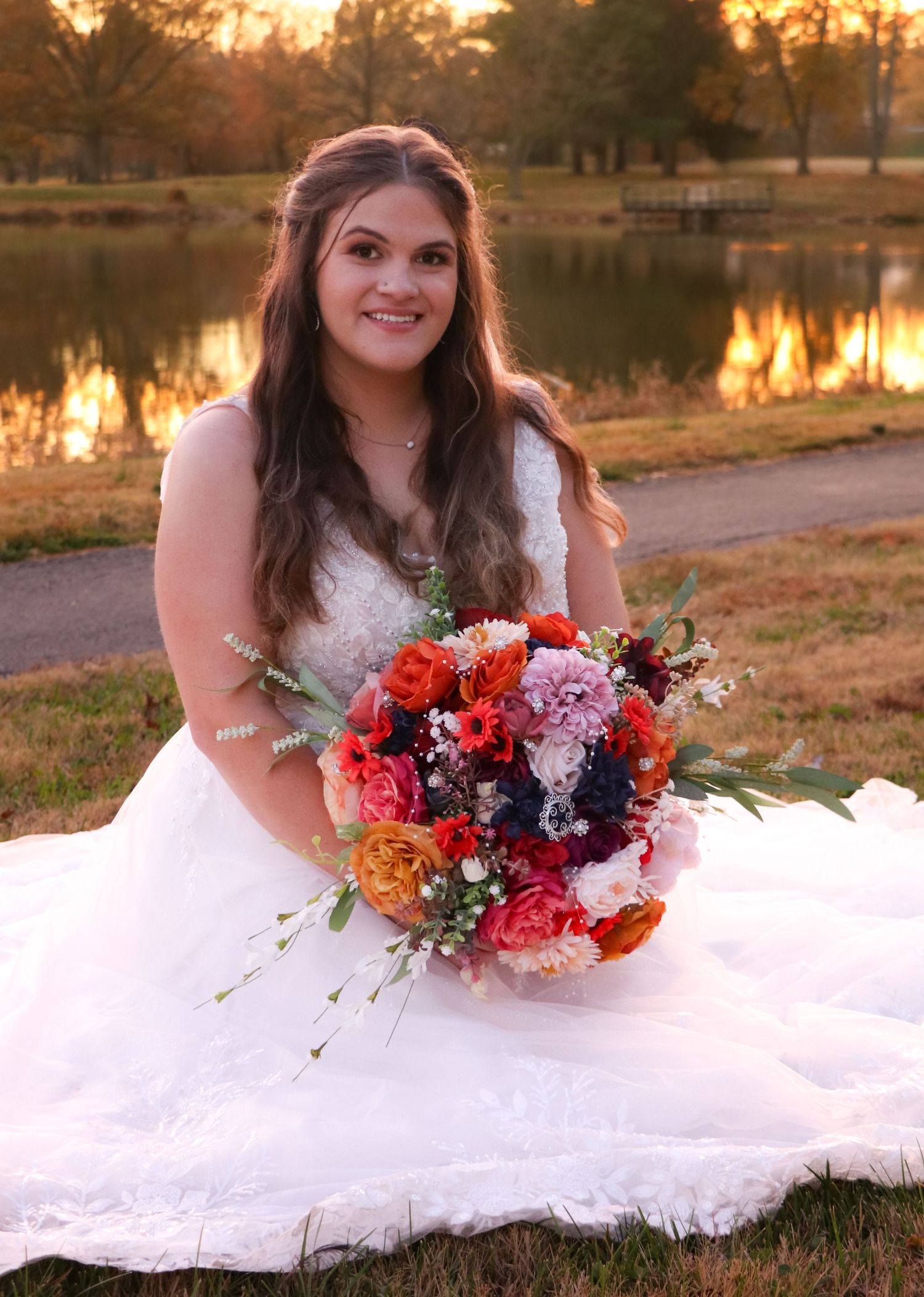 Bride in white dress lying on grass holding vibrant coral and navy floral bouquet at sunset.