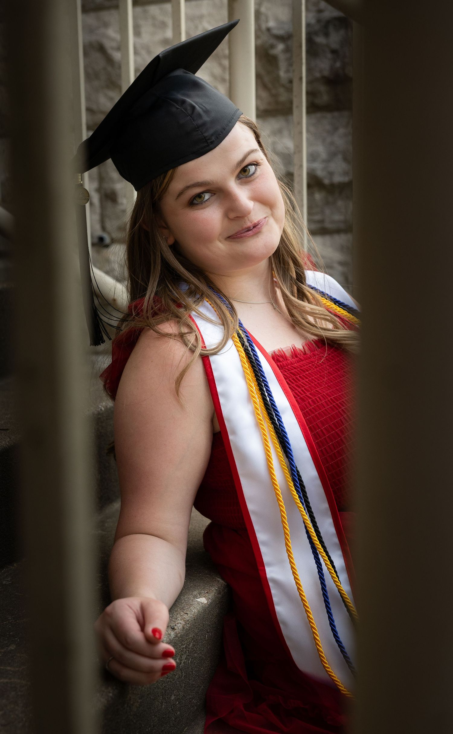 Young woman in graduation cap and red gown with honor cords smiles while leaning on a railing outdoors.