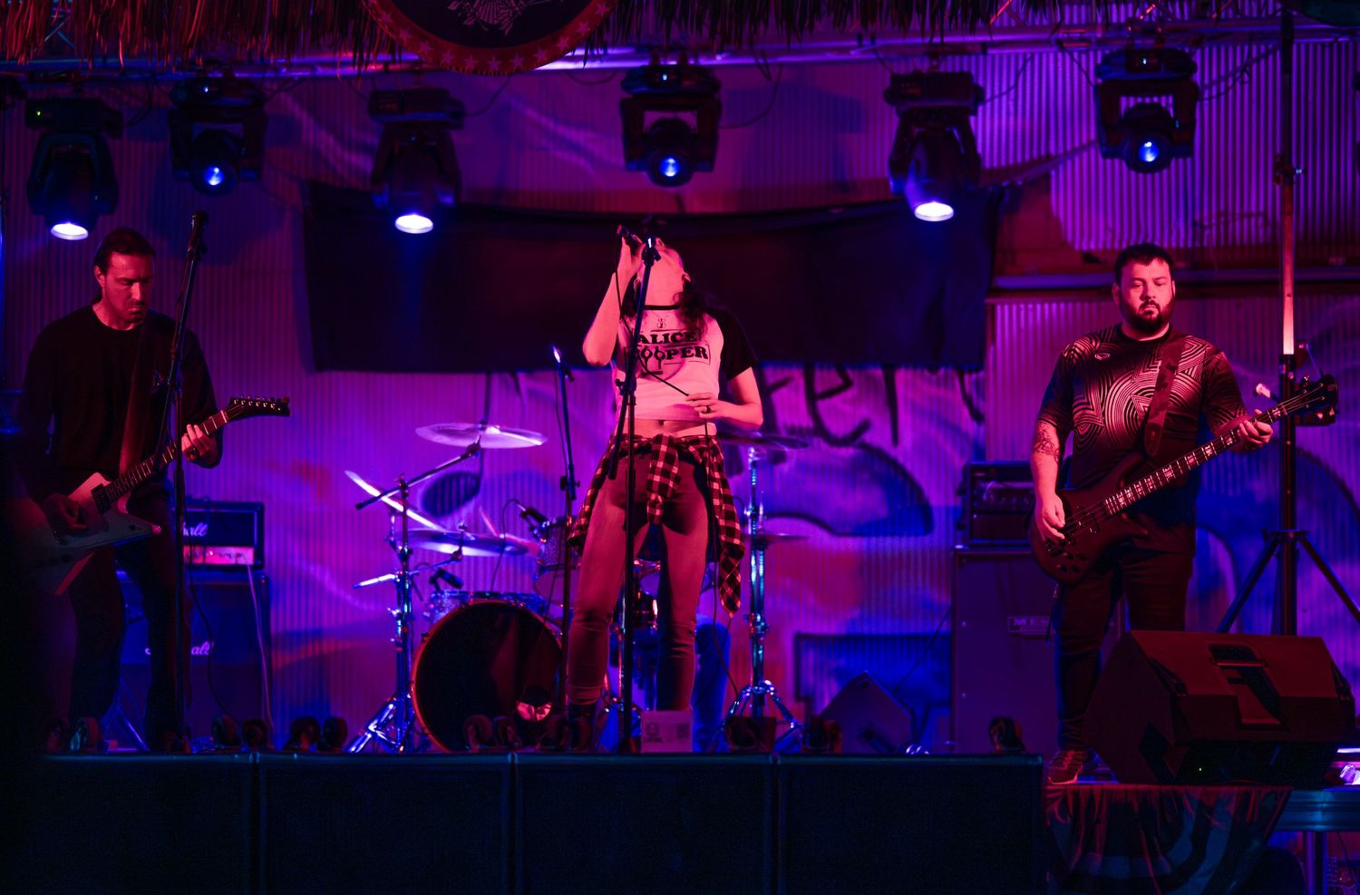 Musicians perform on stage in a dark venue illuminated by vibrant purple lighting during a live concert performance.