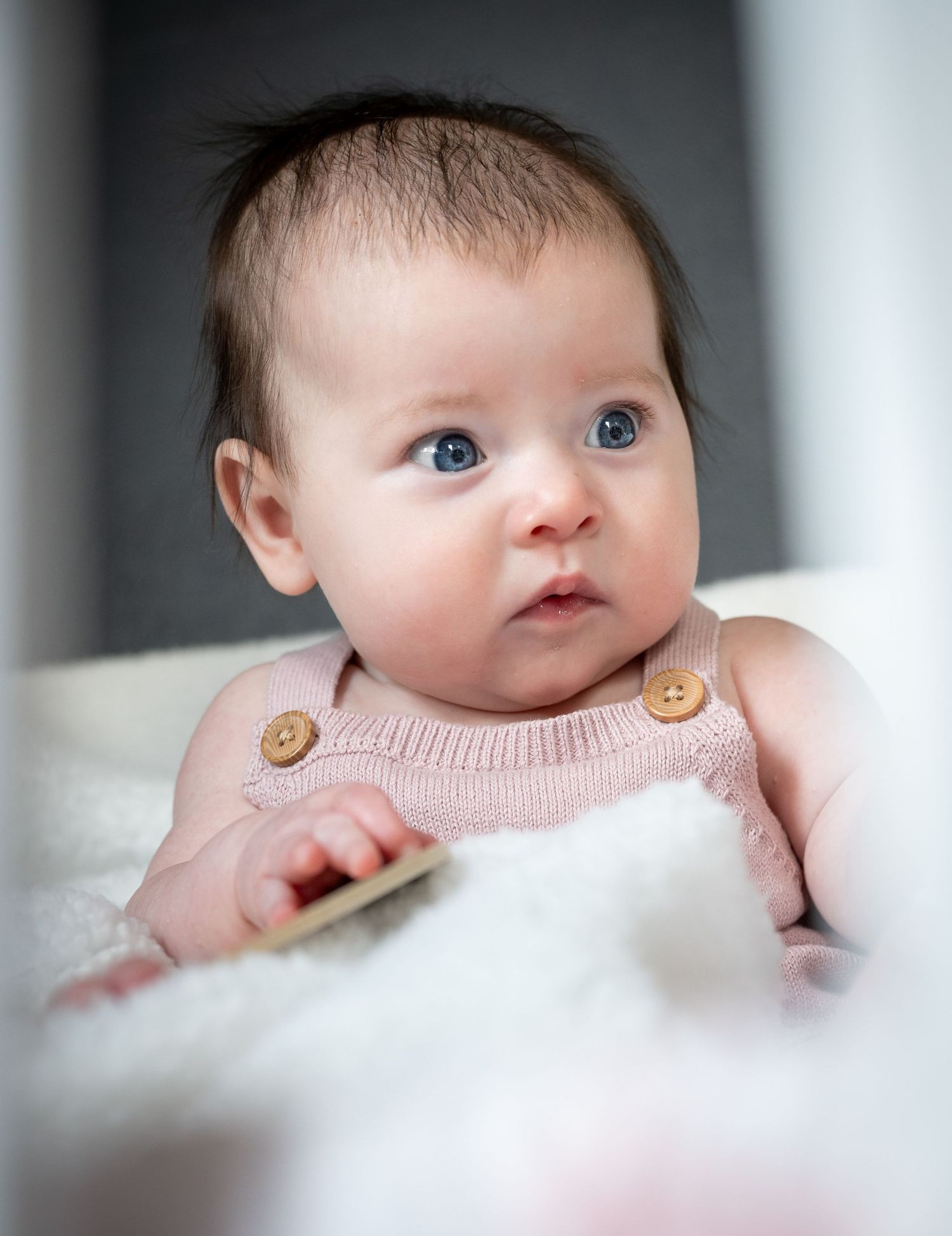 Baby with bright blue eyes peeks over white blanket in soft focus portrait series.