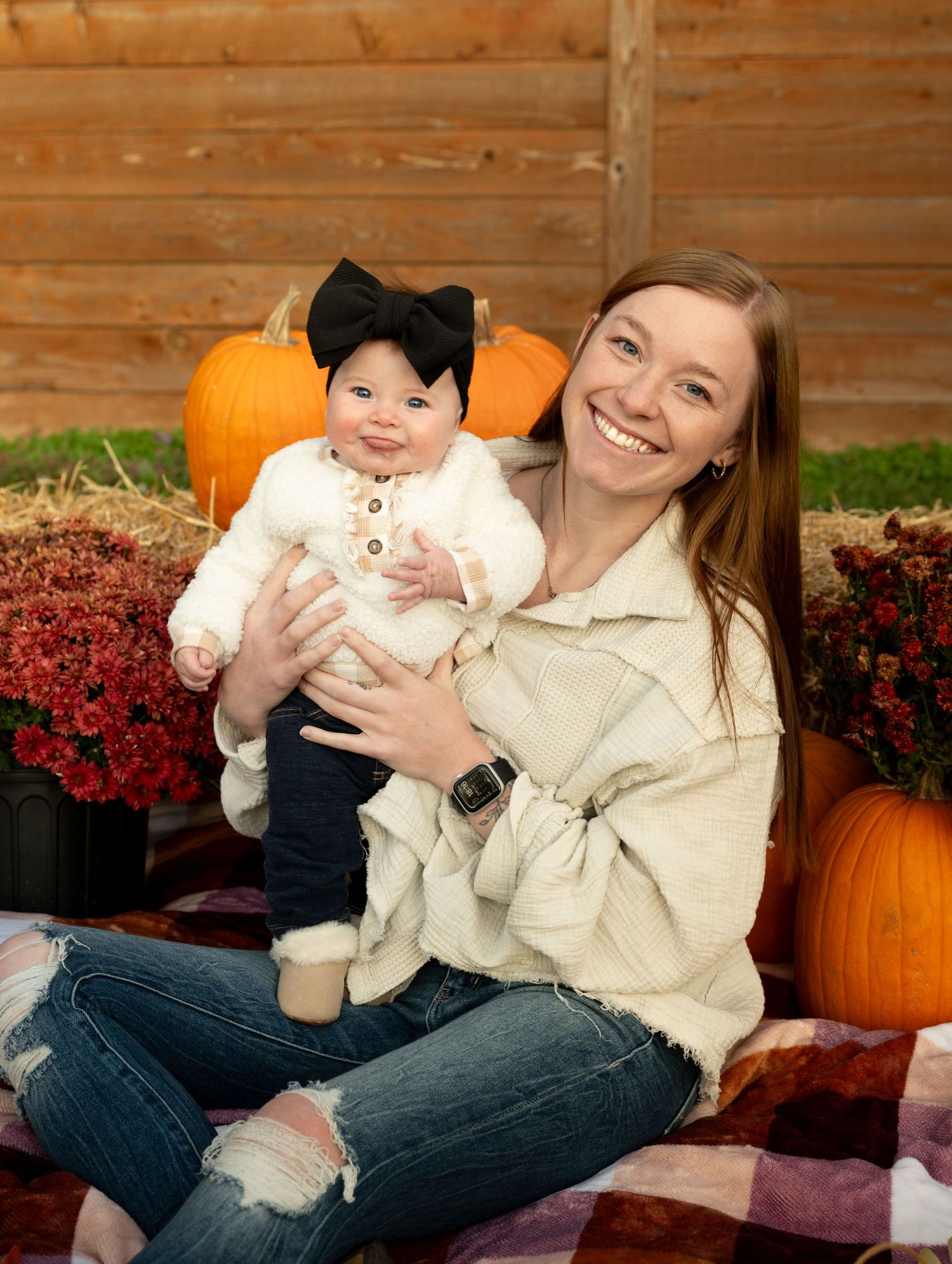 Fall portrait with baby wearing black bow on blanket surrounded by pumpkins and red mums on wooden backdrop.