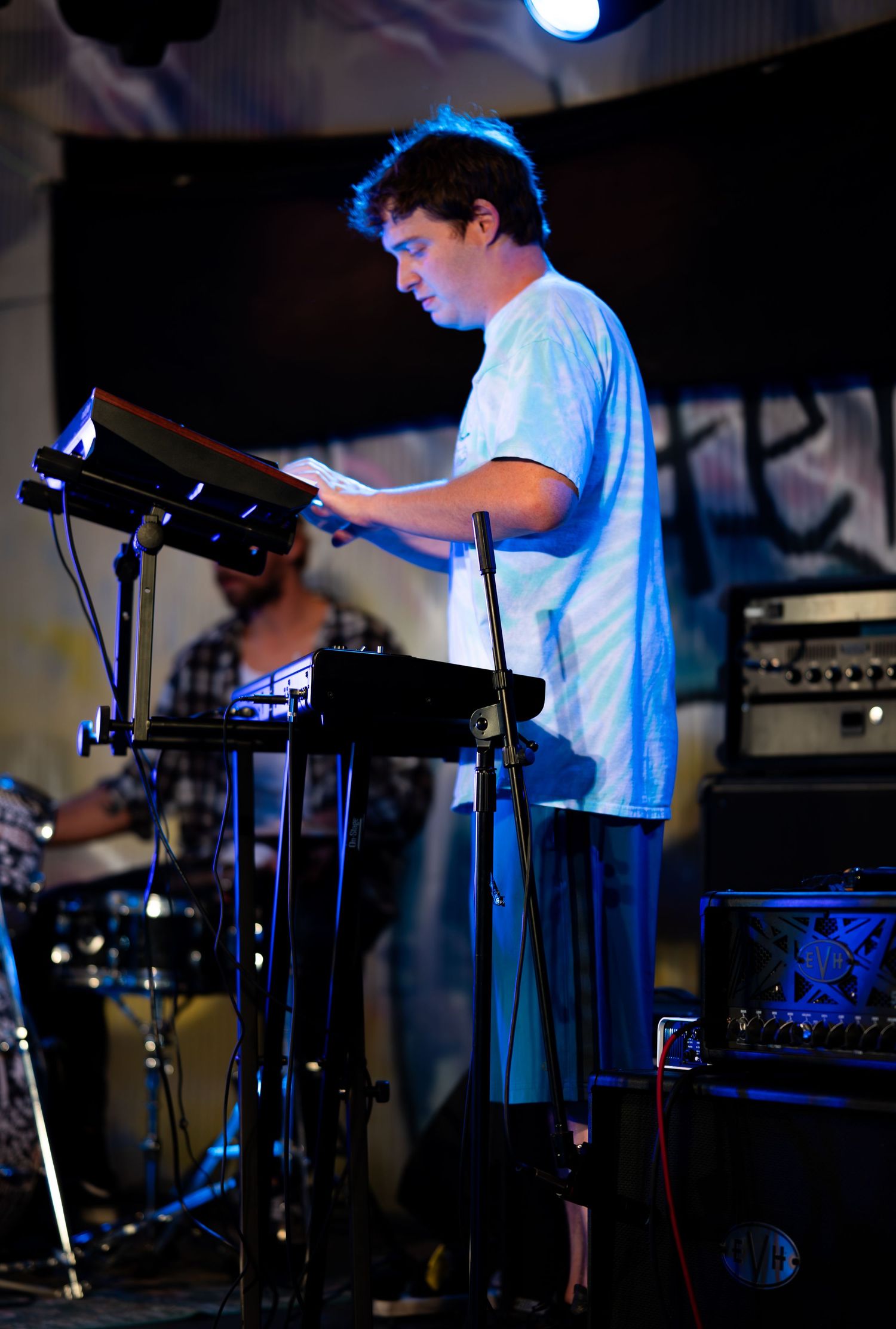 Musician performs on stage with electronic keyboard equipment under blue lighting at a live music venue.