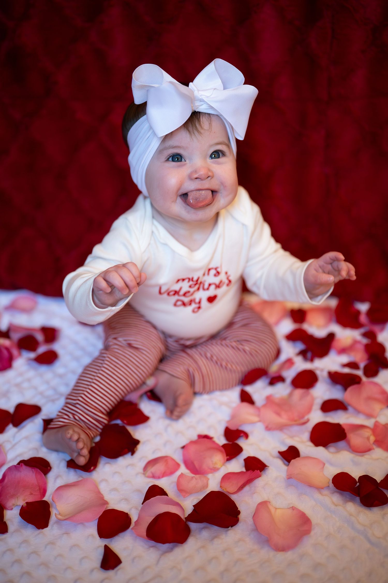 Baby in white outfit with bow lying on white blanket surrounded by pink and red rose petals.