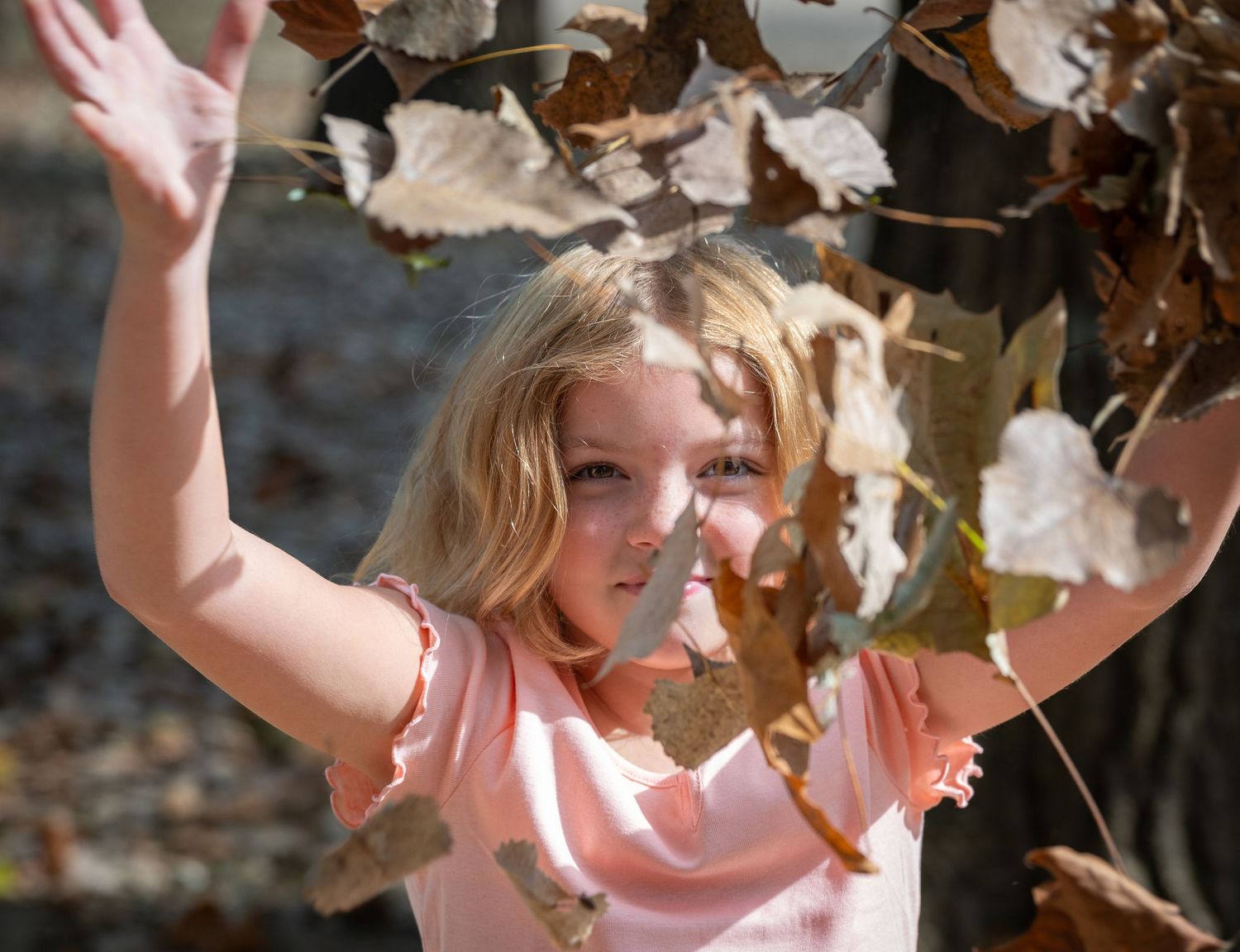 A young person in a pink shirt plays joyfully with falling autumn leaves on a sunny day.