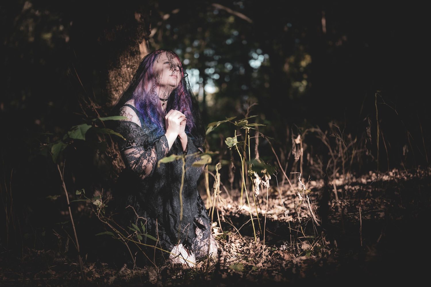 A person in dark clothing sits on fallen leaves in a dimly lit forest setting during golden hour.