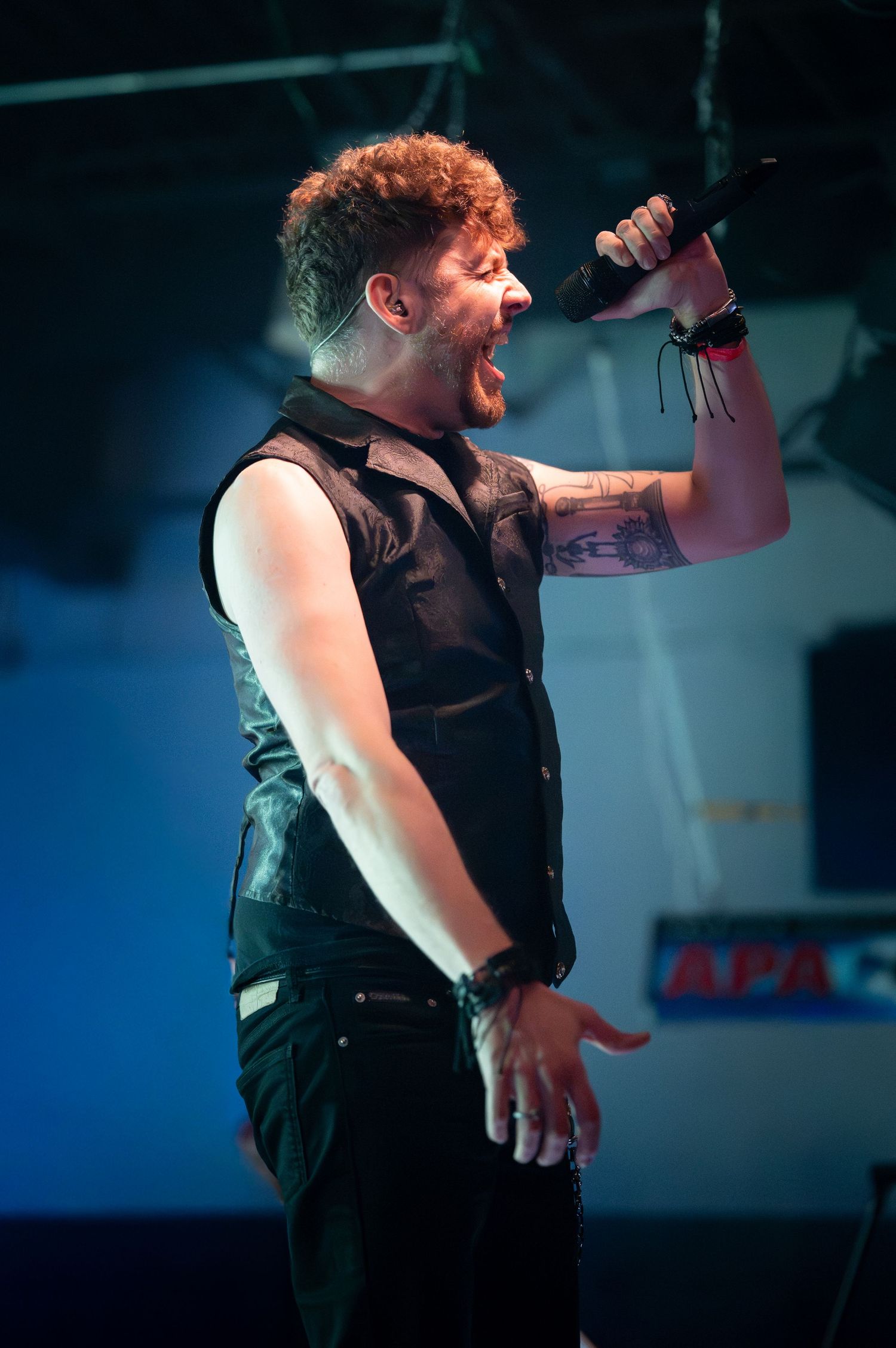 Performer in black sleeveless shirt singing passionately on stage with dramatic blue lighting in background.