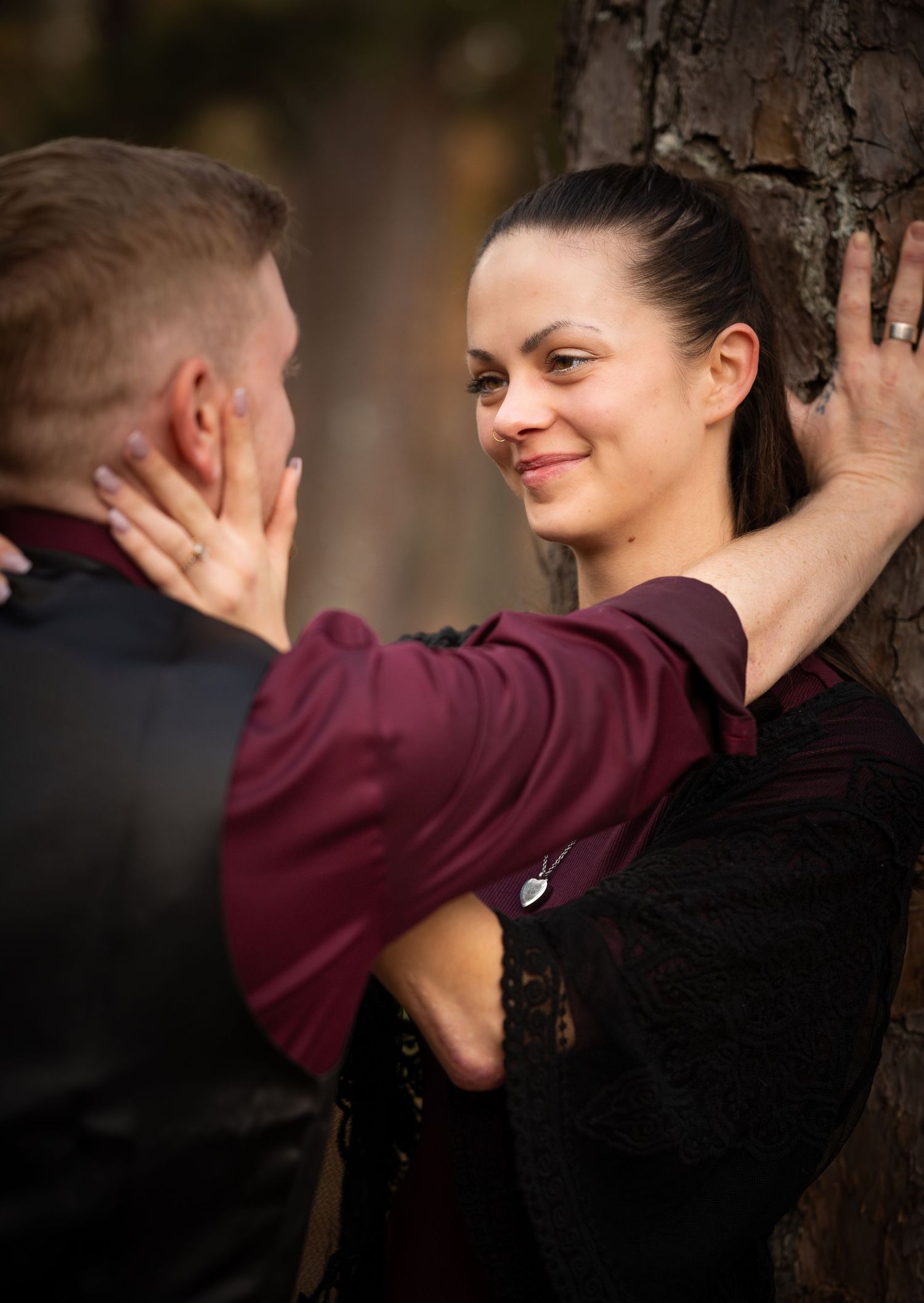 A romantic moment captured as a couple shares an intimate exchange against a tree trunk in burgundy and black attire.