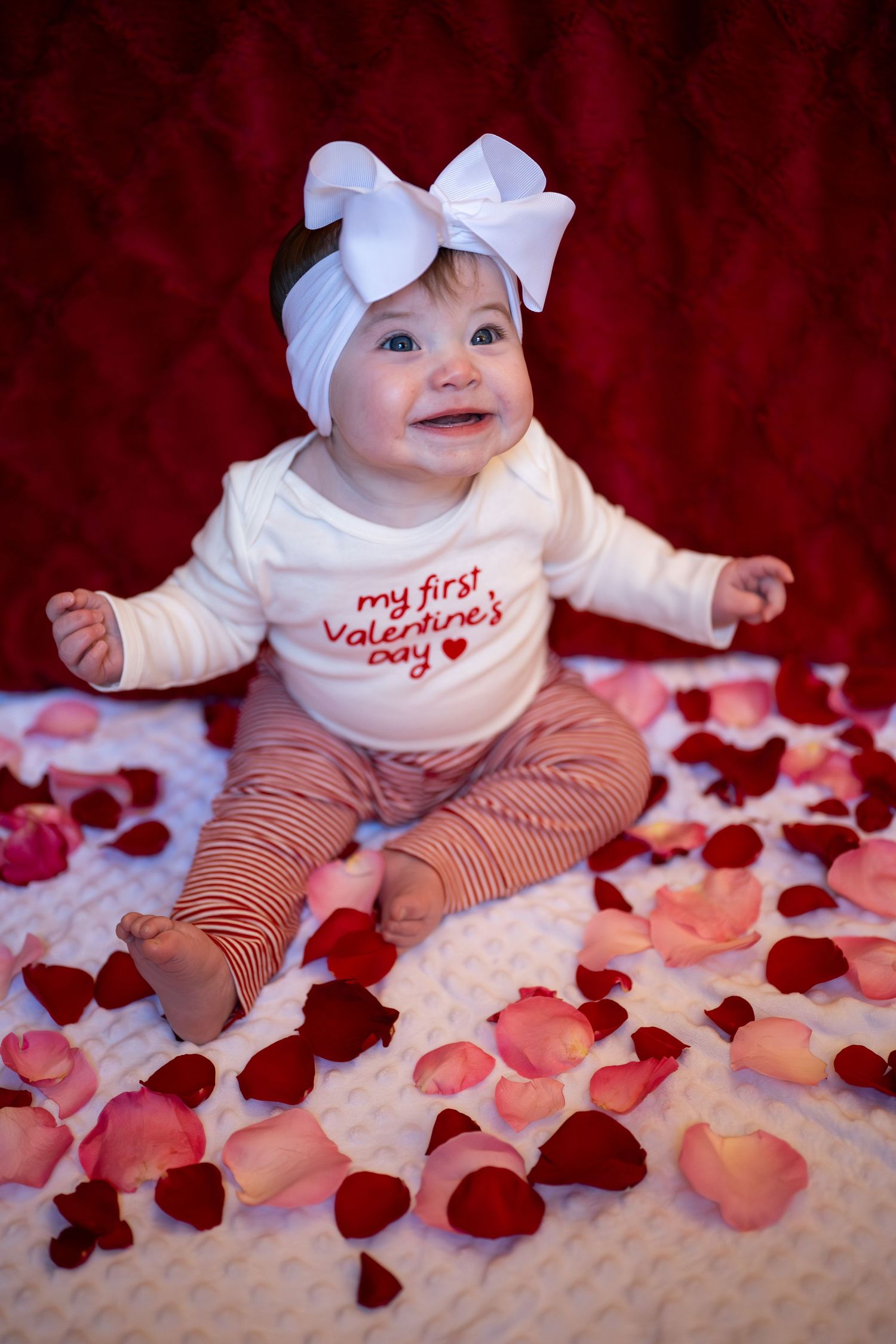 Baby wearing white bow and Valentine's outfit sits on blanket surrounded by scattered pink and red rose petals.
