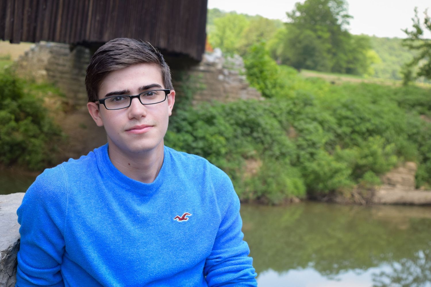 Person wearing glasses and a blue sweater sits outdoors near a scenic pond with trees in the background.