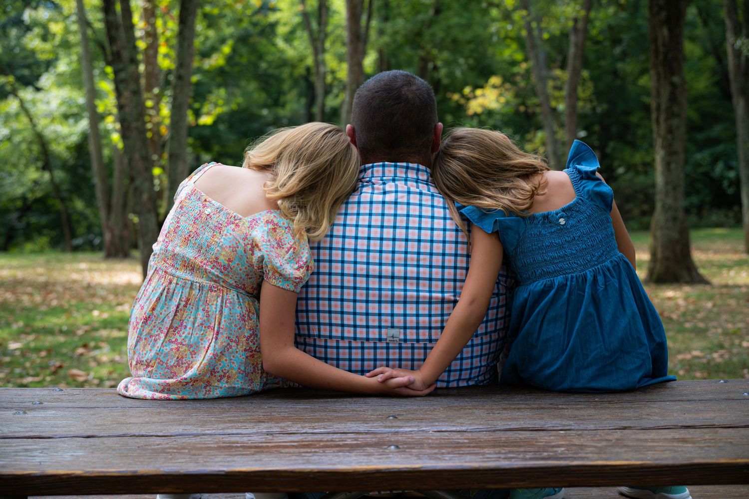 Two people sit close together on a wooden bench in a peaceful park setting surrounded by green trees.