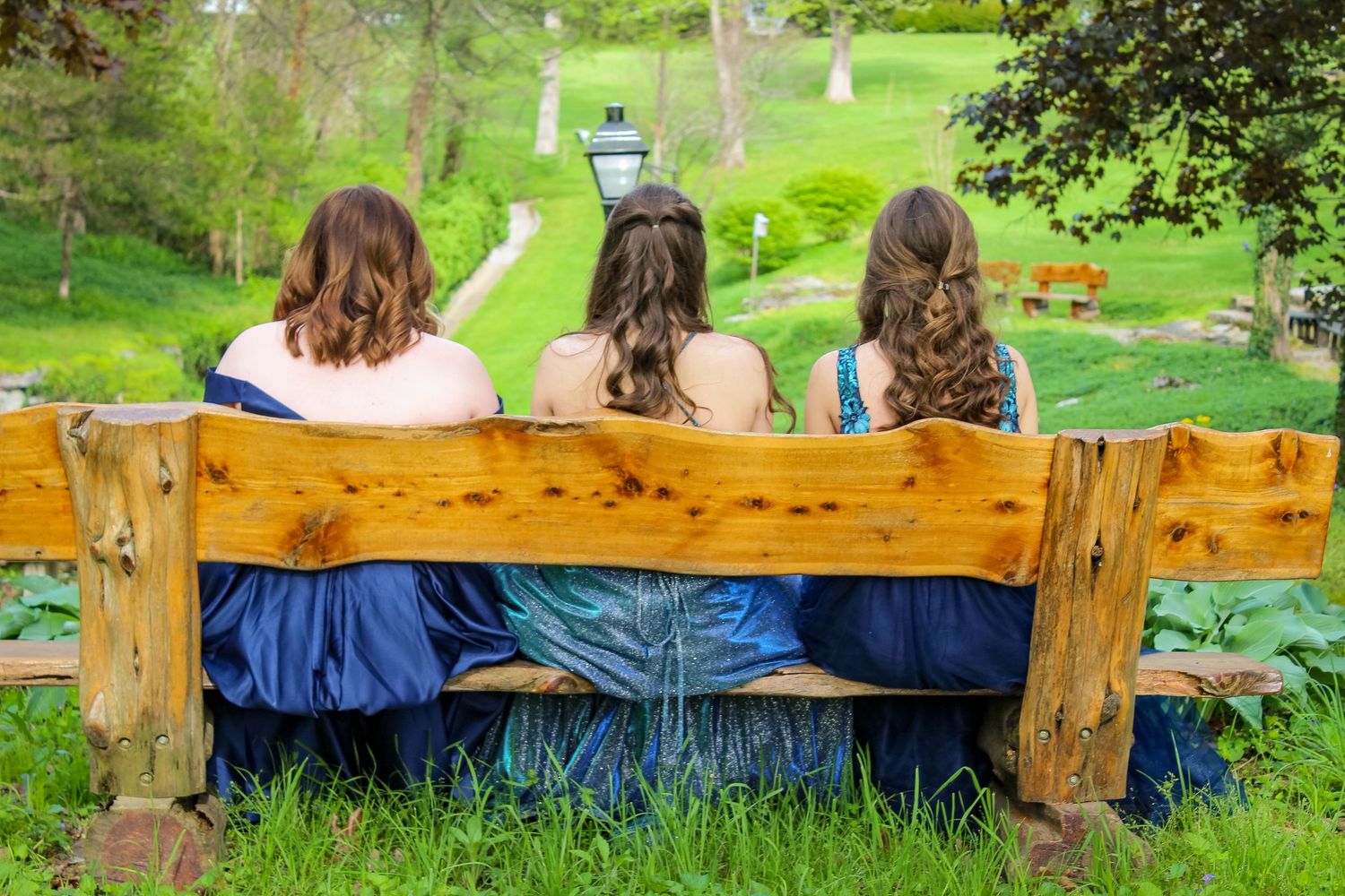 Three people in blue dresses sit together on a rustic wooden bench overlooking a scenic green hillside path.