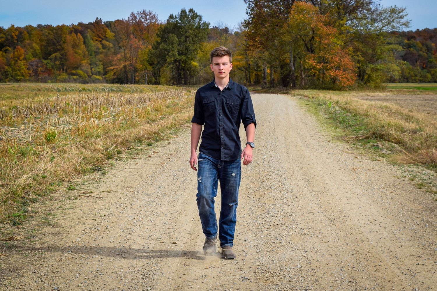 A person walks along a winding dirt road through autumn countryside on a sunny day.