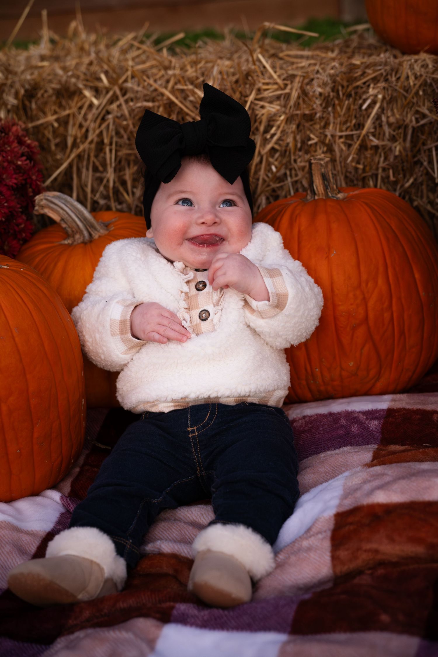 A baby in a white sweater sits among autumn pumpkins on a brown blanket in a fall photo shoot setting.