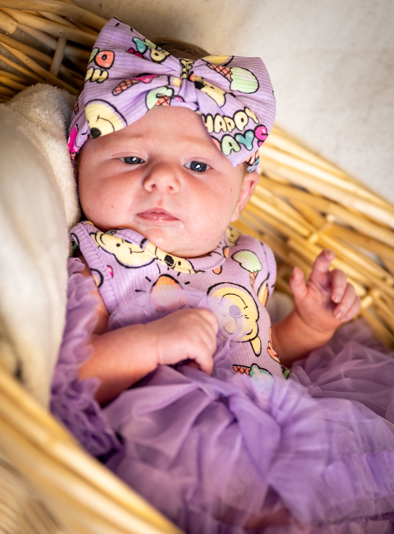 A baby wearing a purple onesie with unicorn patterns lies on a light colored surface.