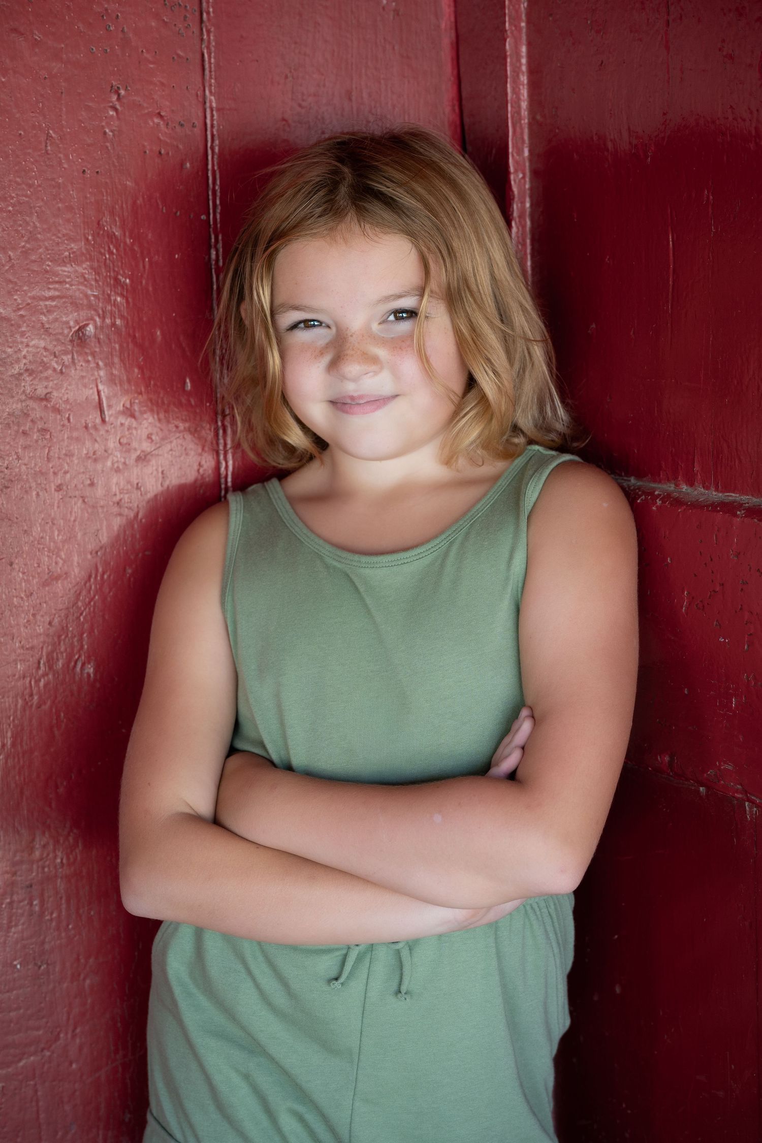 Young person in a sage green tank top poses with crossed arms against a dark red wall.