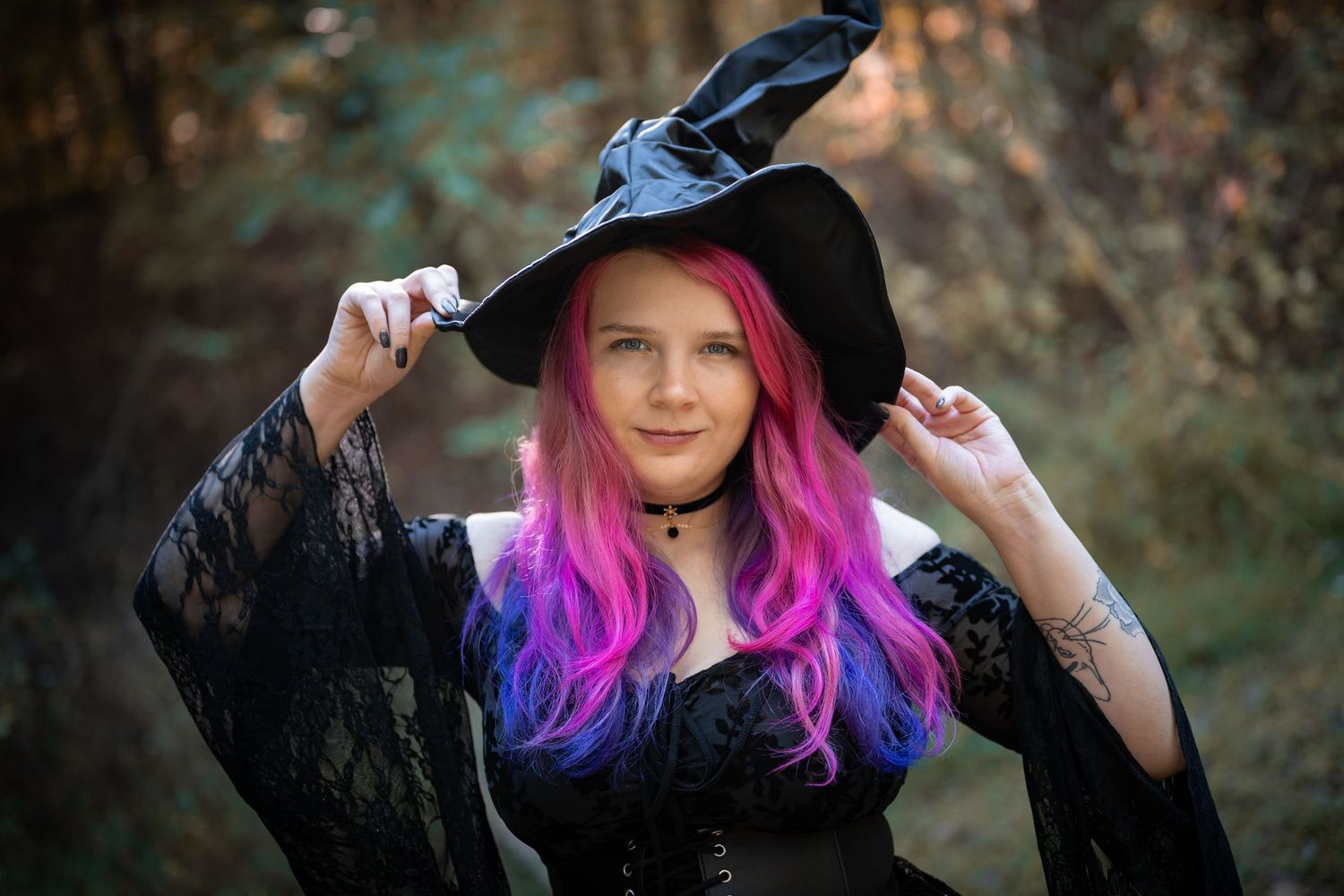 Young witch with vibrant pink and purple hair wearing a black hat and dress in a magical forest setting.