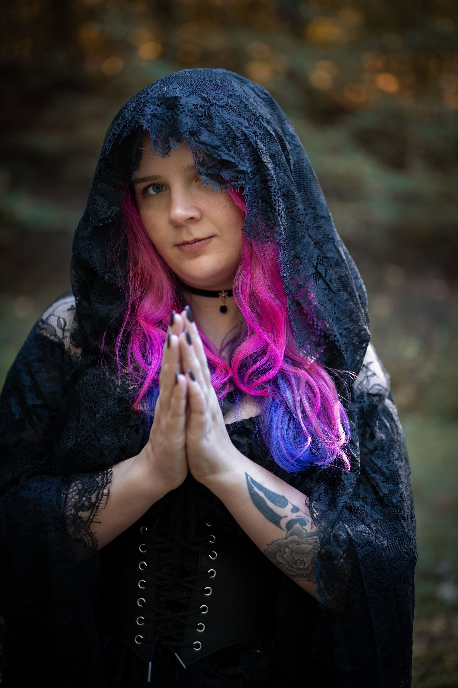 A gothic figure in black lace veil with vibrant pink and purple hair holds their hands in prayer pose outdoors.