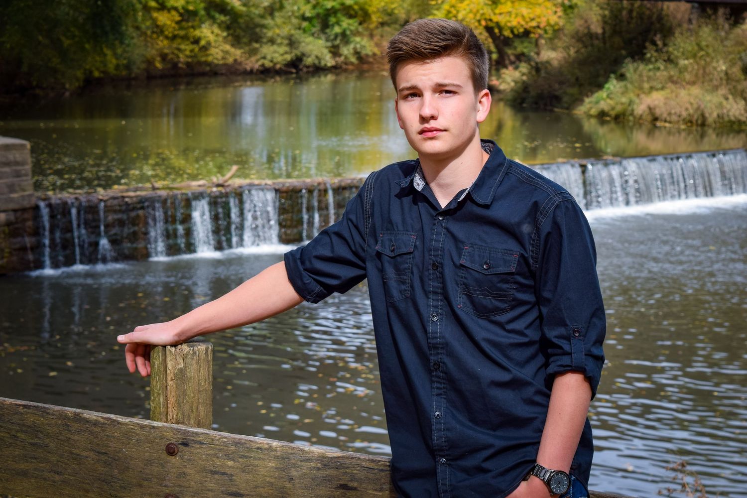 A person in a navy blue shirt stands by a wooden railing near a scenic waterfall and river.