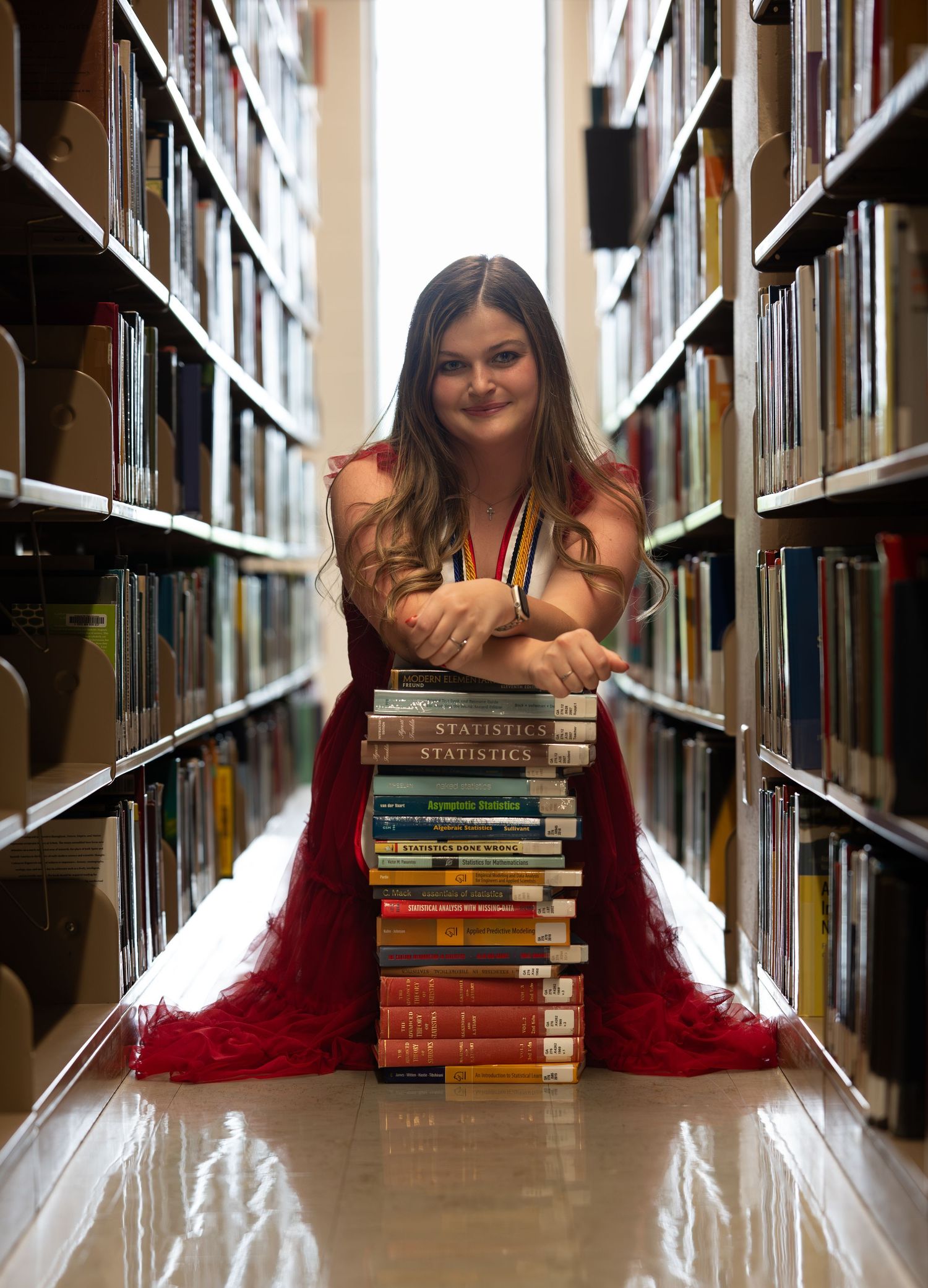 Young woman in red dress poses with stacked books in library aisle for senior portrait photo.