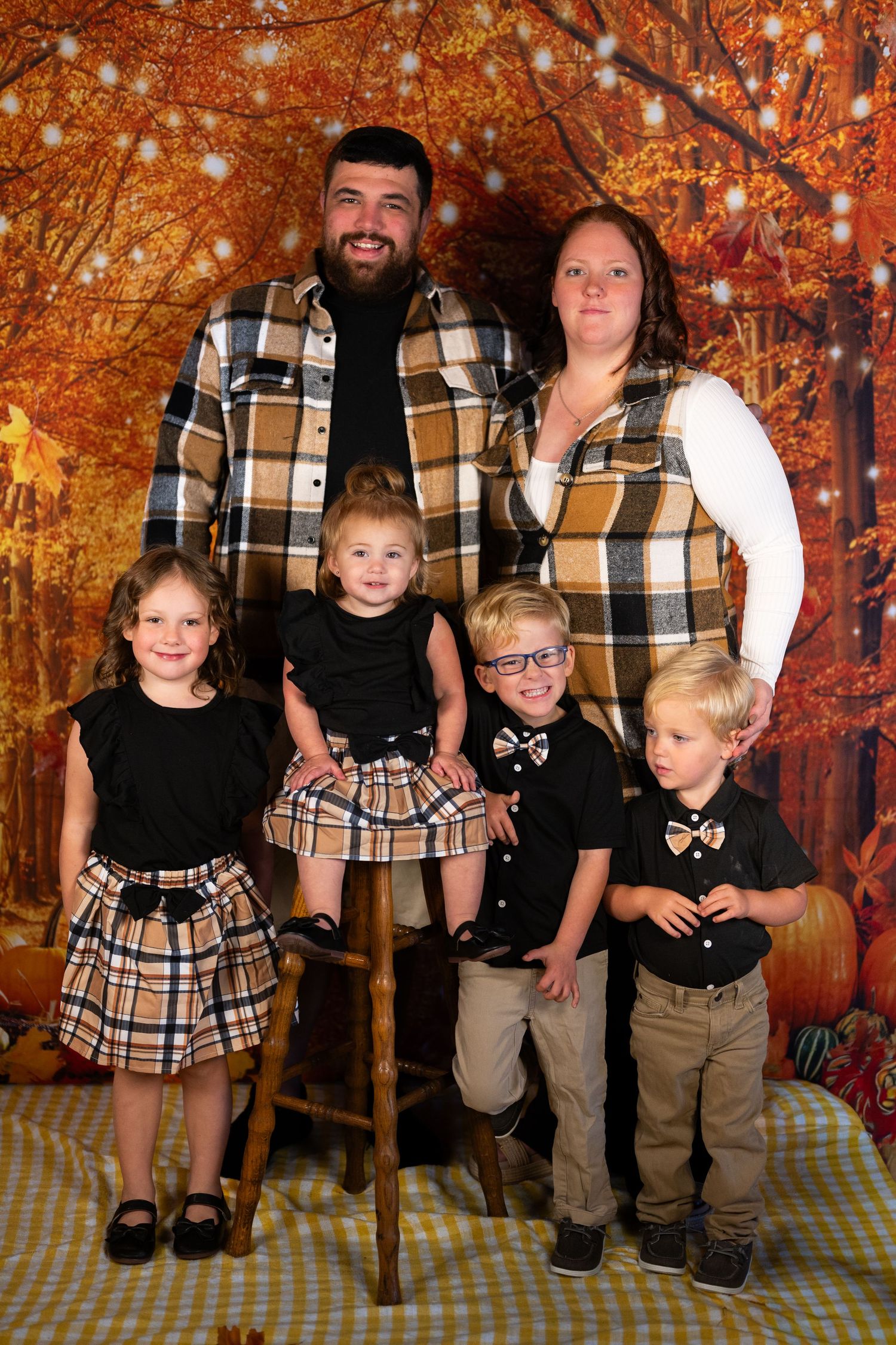 A family of six poses in coordinating plaid outfits against a warm autumn backdrop with pumpkins.