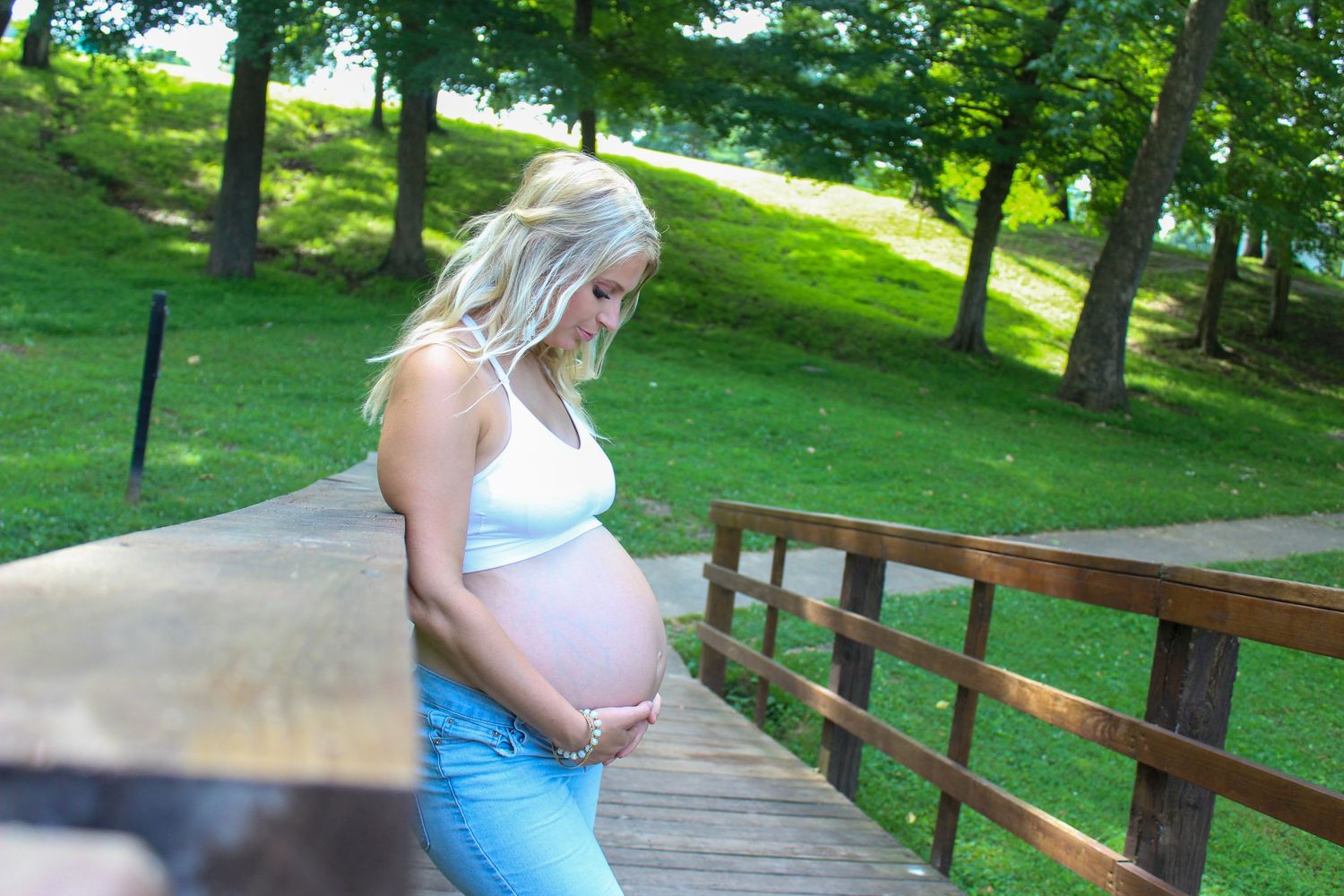 A pregnant woman in a light blue dress walks along a wooden bridge pathway through a lush green park.
