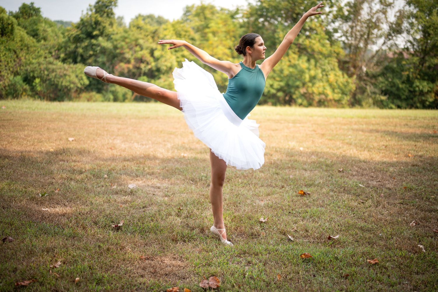 Ballet dancer performs an arabesque pose in a white tutu on a grassy field during golden hour.
