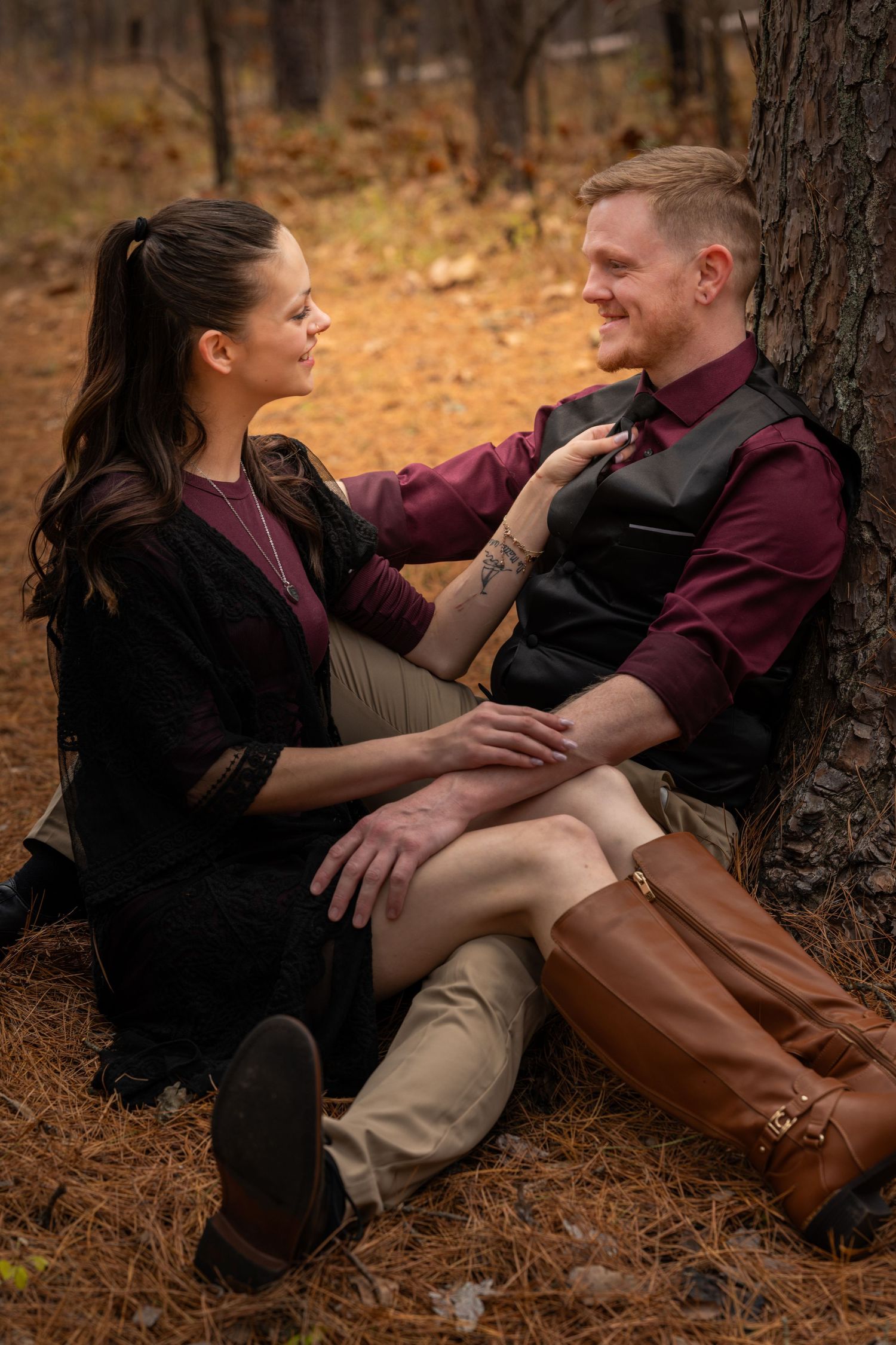 A couple in burgundy and black clothing share intimate moments while sitting against a tree in an autumn forest setting.