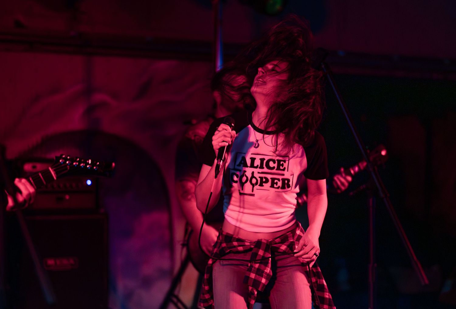 Musicians perform on stage in a dark venue illuminated by purple and red lighting during a live concert.