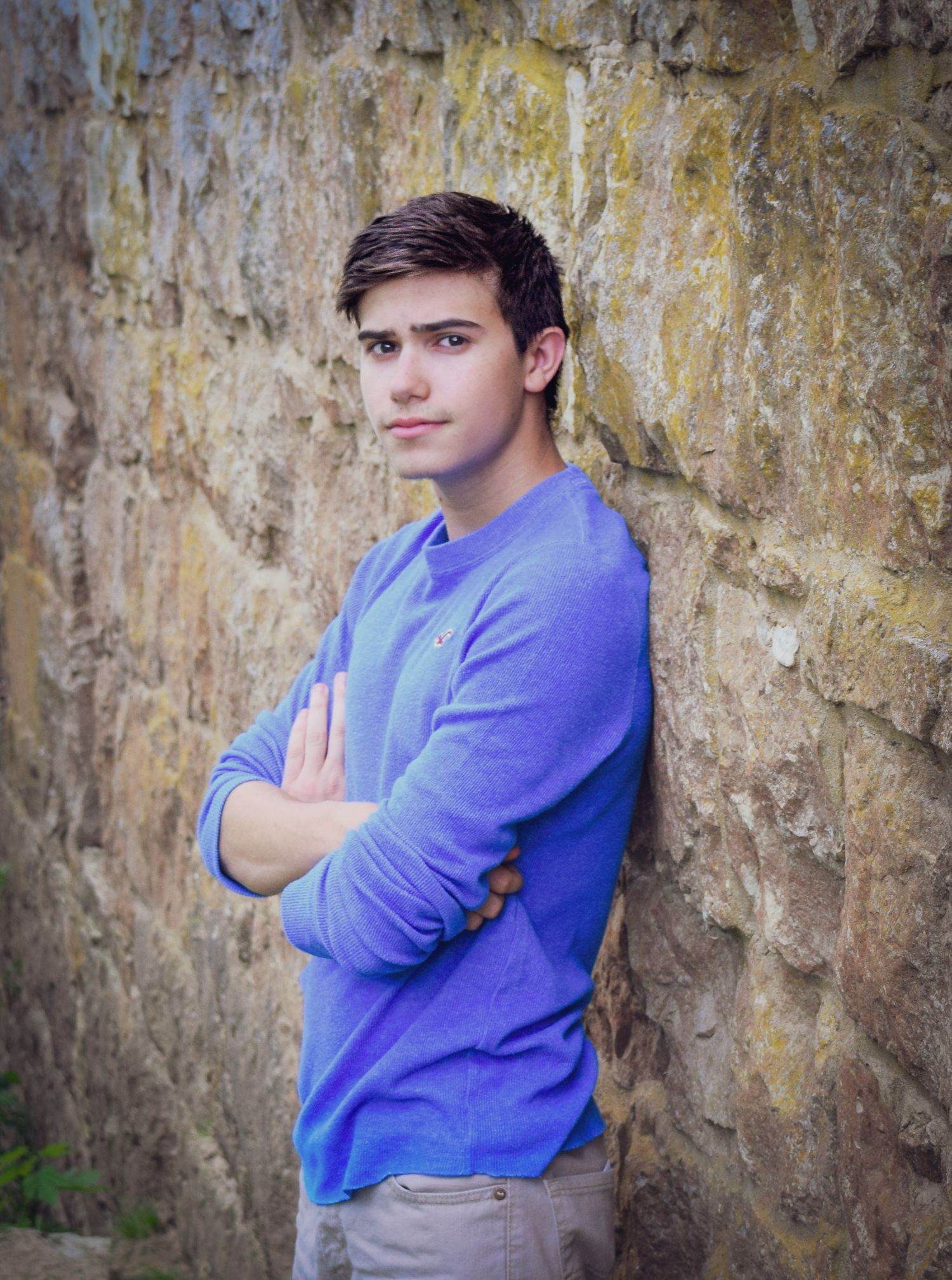 A person in a purple sweater poses against a textured stone wall with arms crossed.