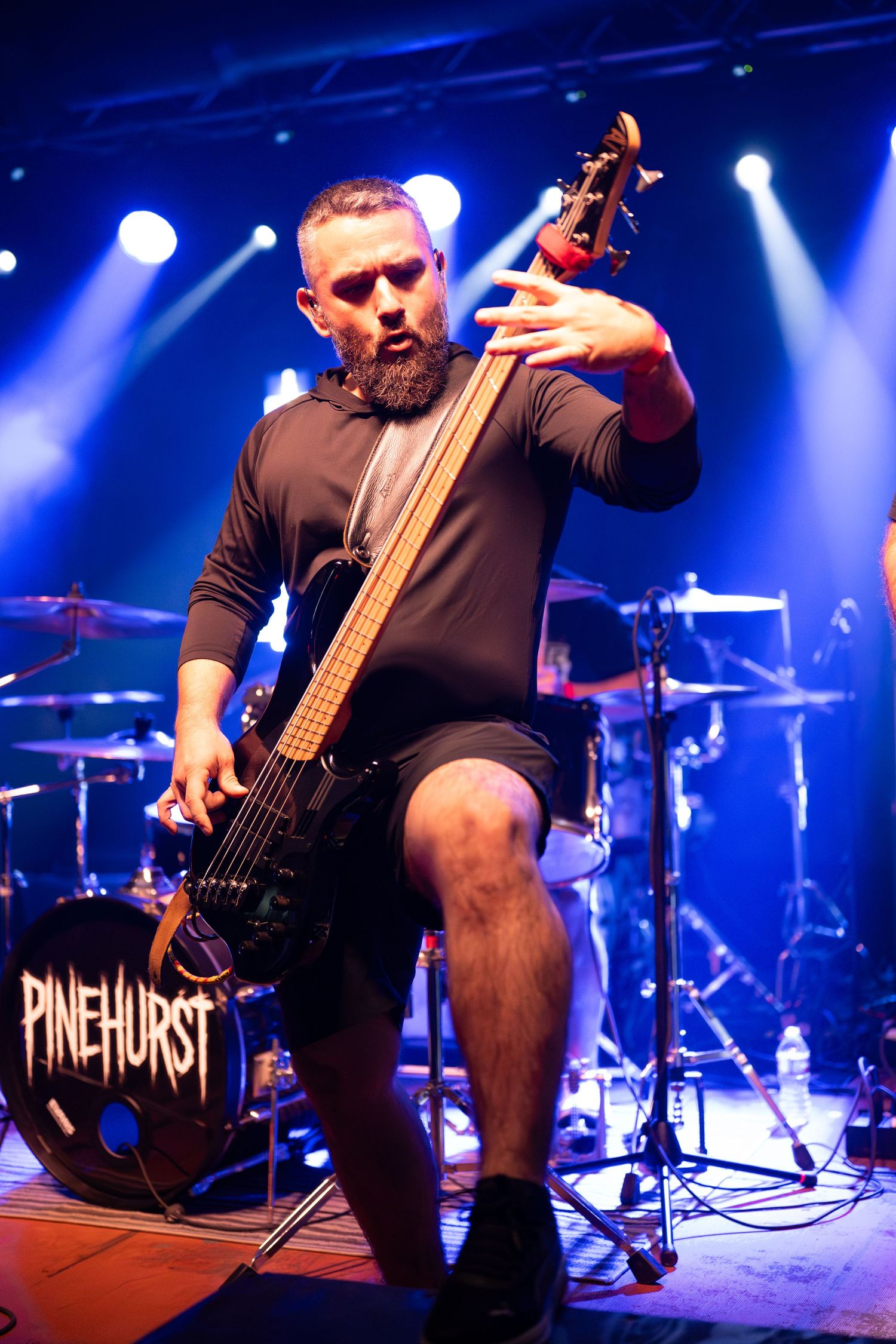 Bass guitarist performs on stage with Pinehurst band logo visible under intense blue concert lighting.