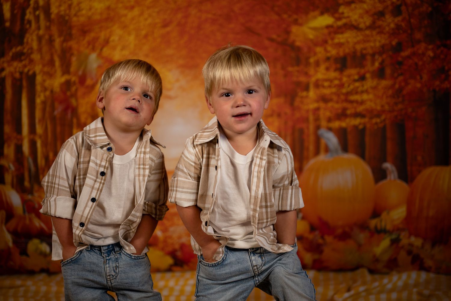 Two small children in matching plaid shirts and jeans pose against an autumn scene with pumpkins.