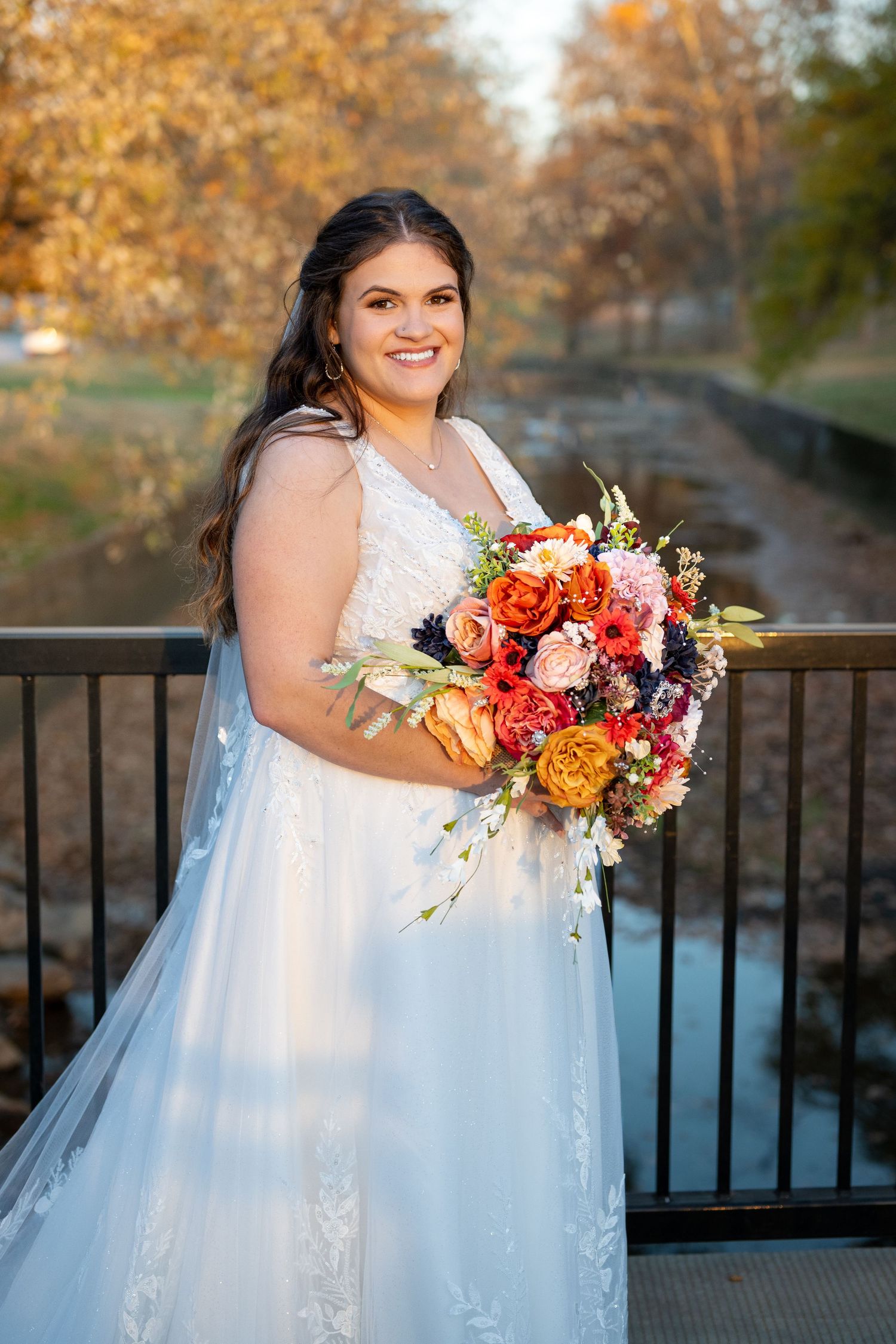 Bride in lace gown holds vibrant fall-colored bouquet against autumn scenery.