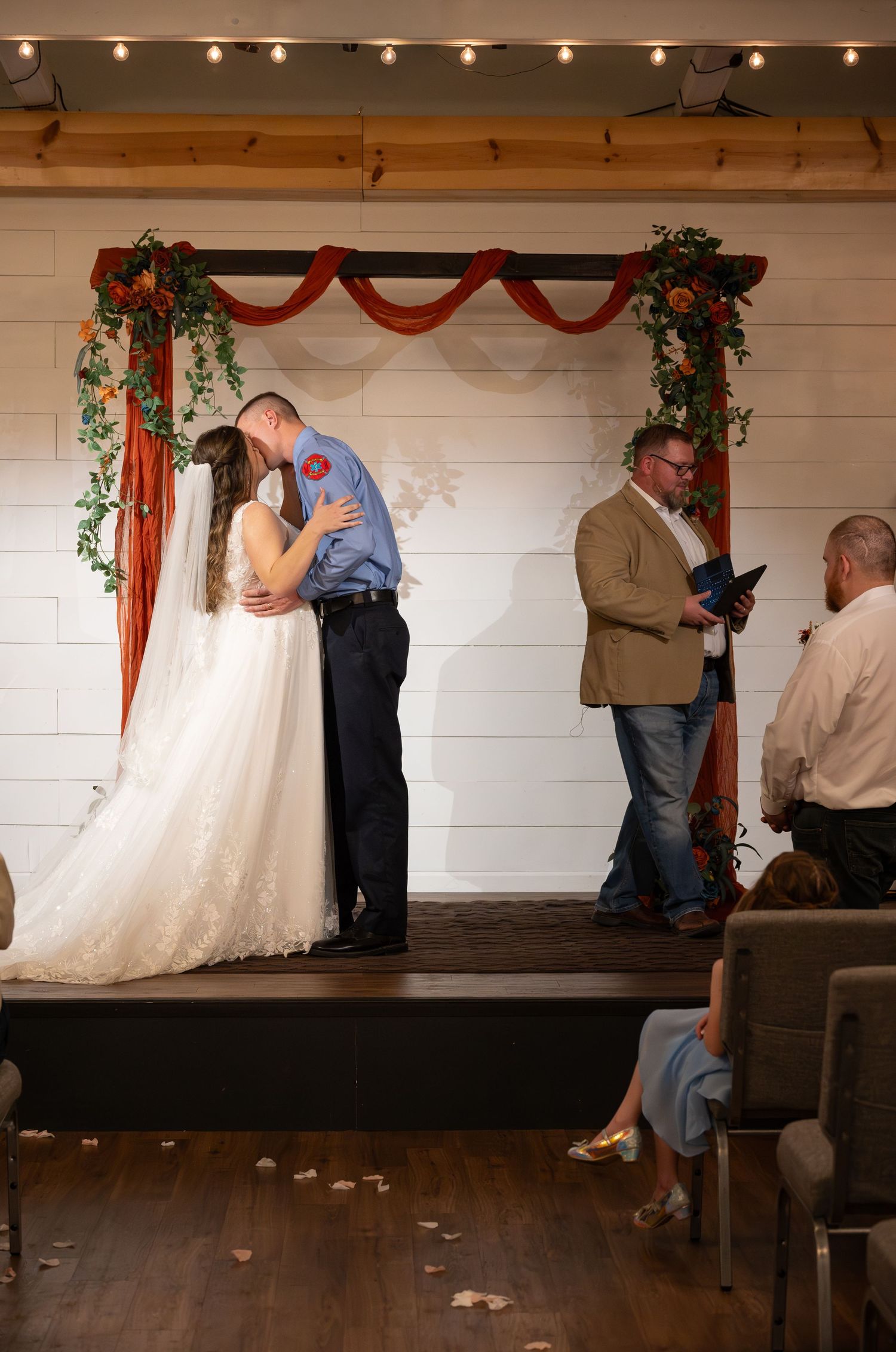 Wedding ceremony on elevated wooden stage with white shiplap walls and decorative draping.