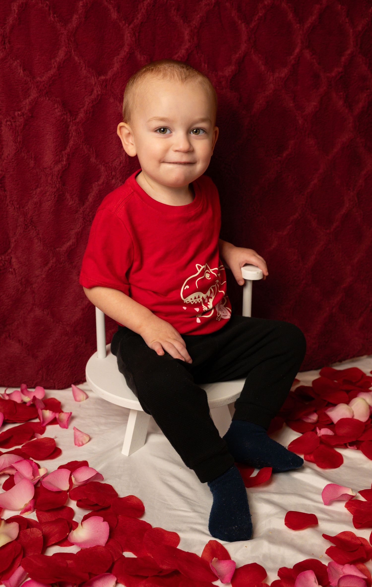 A small child in a red shirt sits on a white surface scattered with red and pink rose petals.