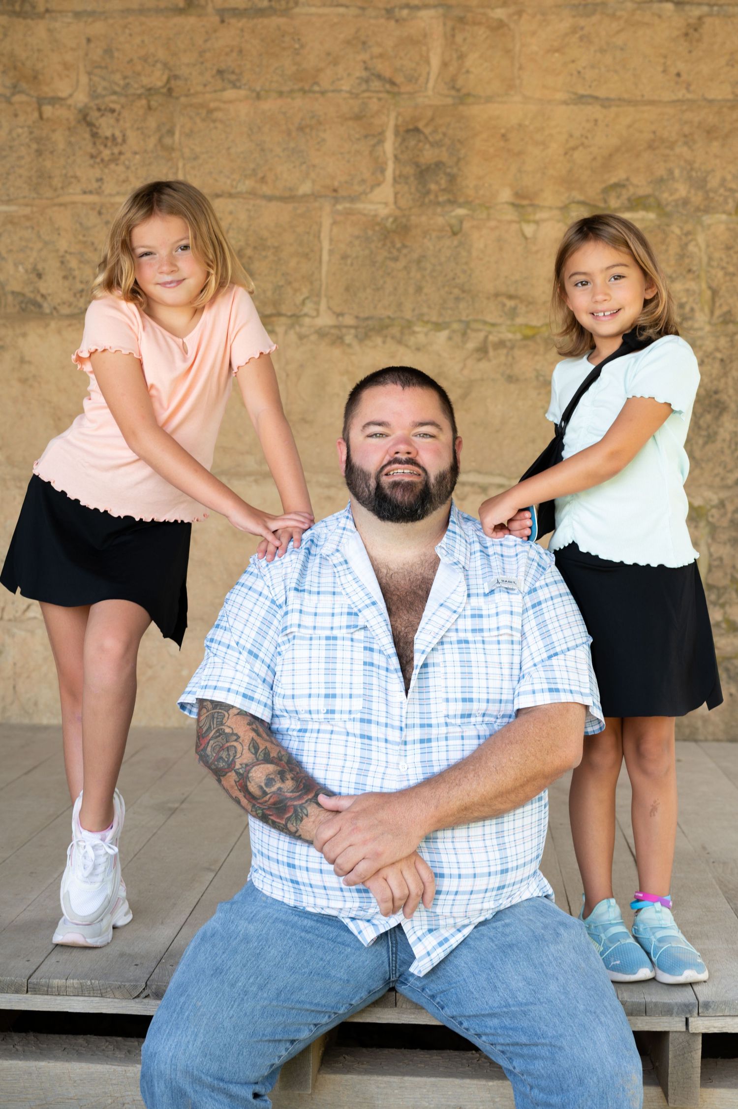 Three people pose together on stone steps with one seated in the center wearing a plaid shirt and jeans.