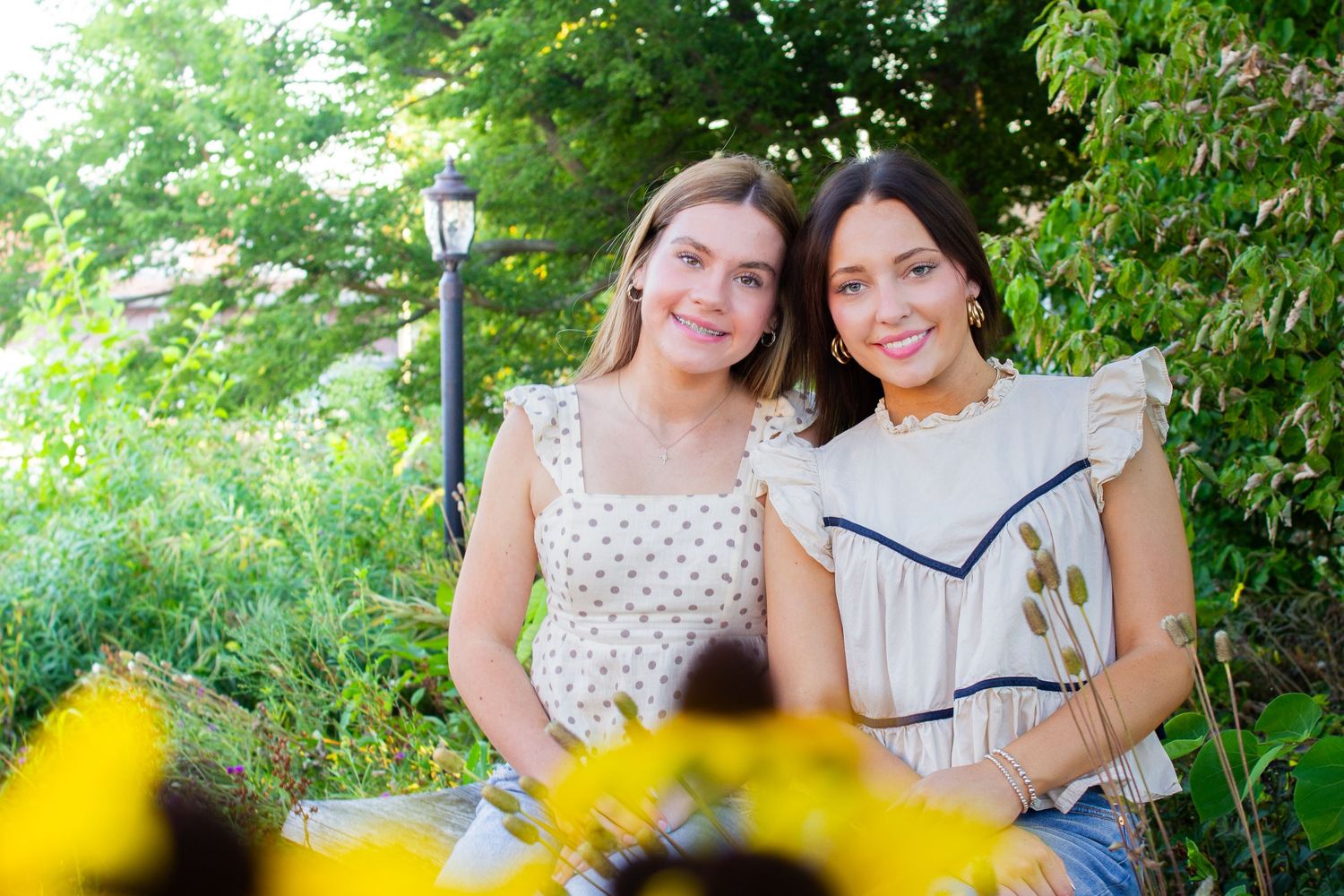 Two people sit together outdoors in a garden setting with green foliage and yellow flowers in the background.
