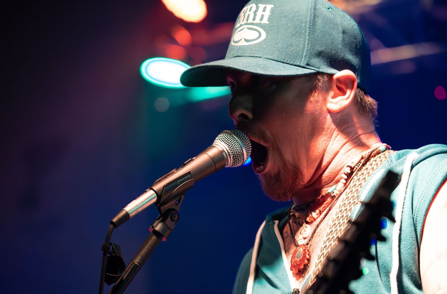 Musician performs on stage wearing a baseball cap under colorful stage lighting at a live concert venue.