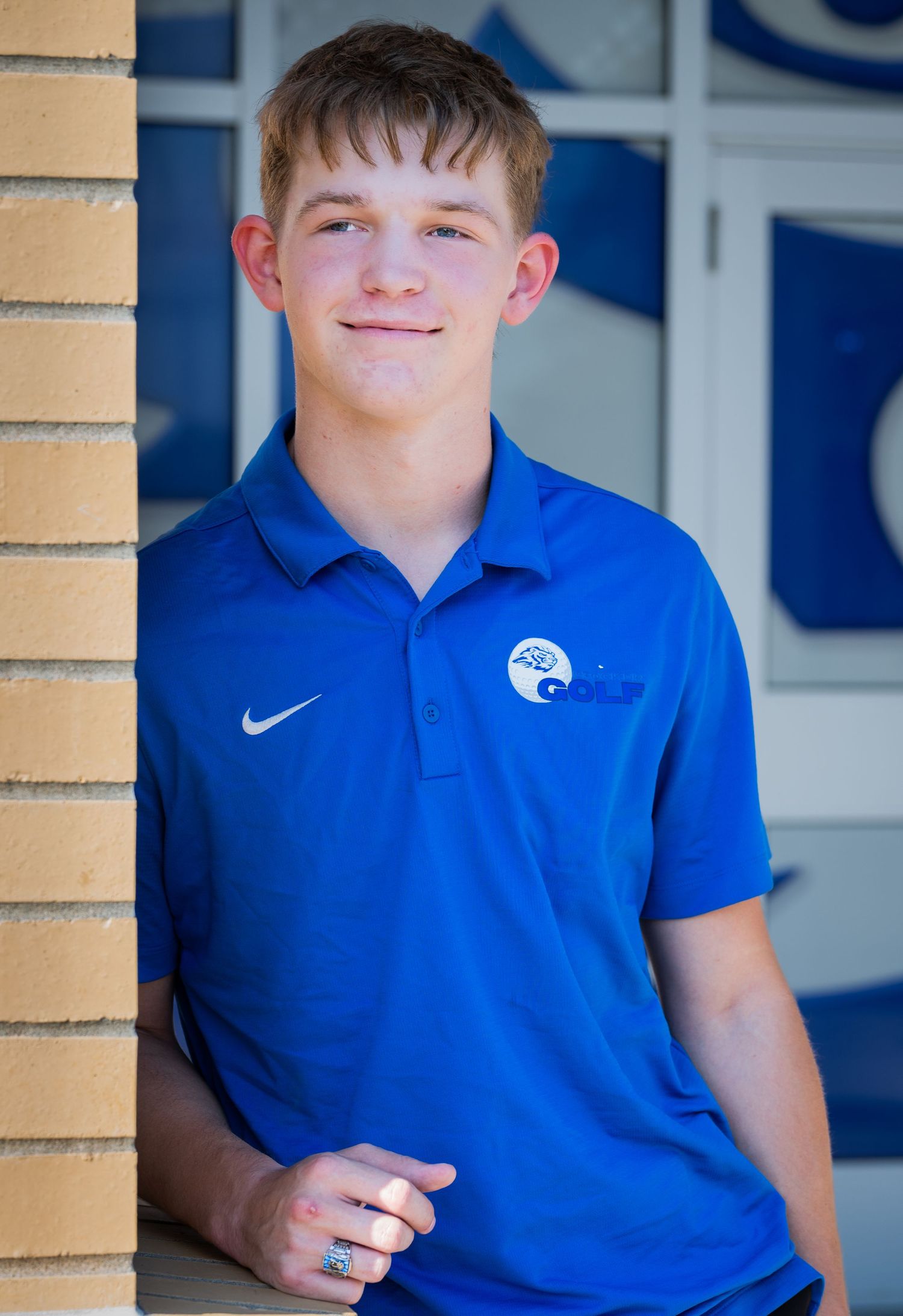 Student in royal blue Nike polo shirt sits casually against beige brick column at school entrance.