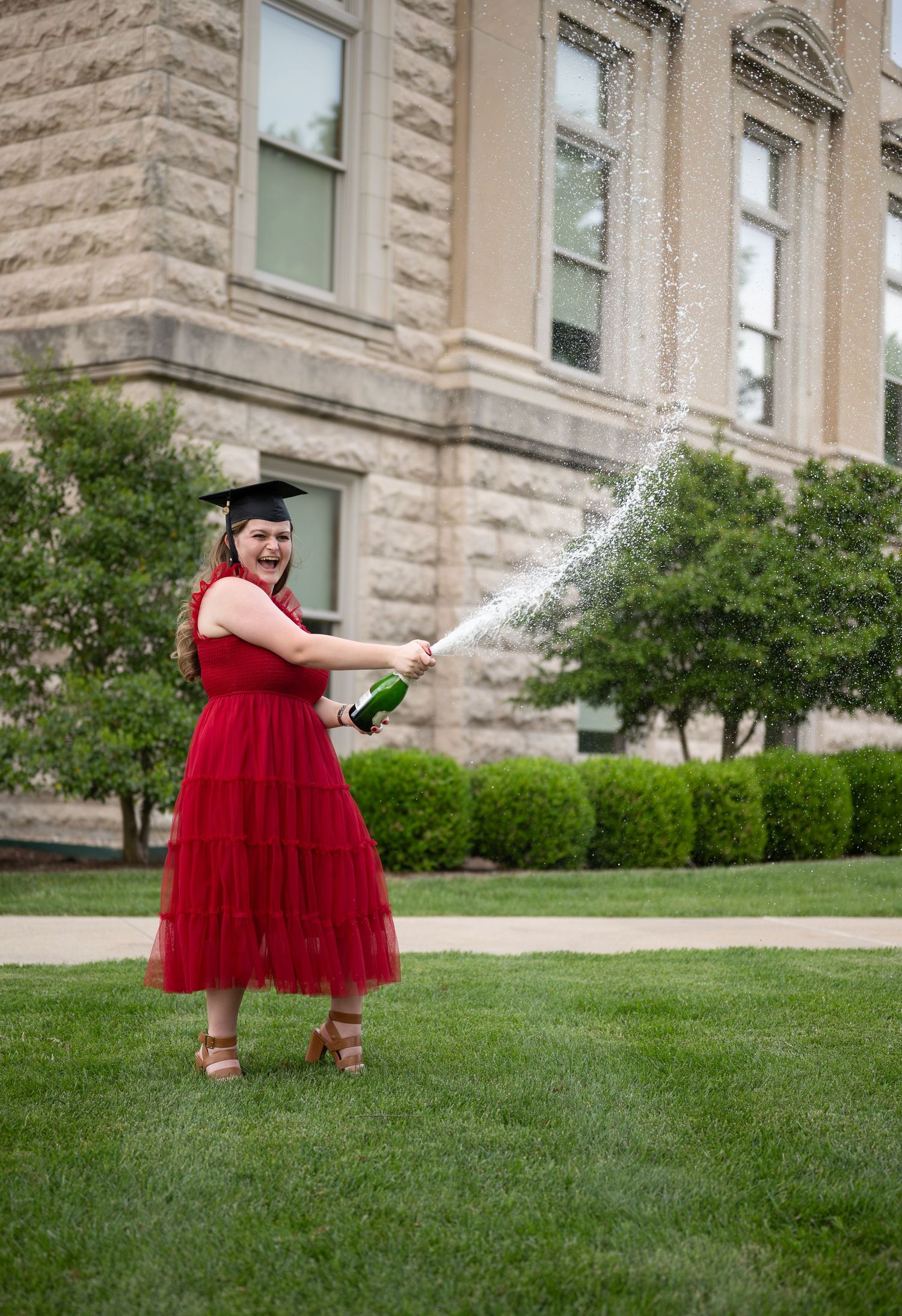A joyful graduate in a red dress and cap sprays champagne outdoors in front of a stone building.