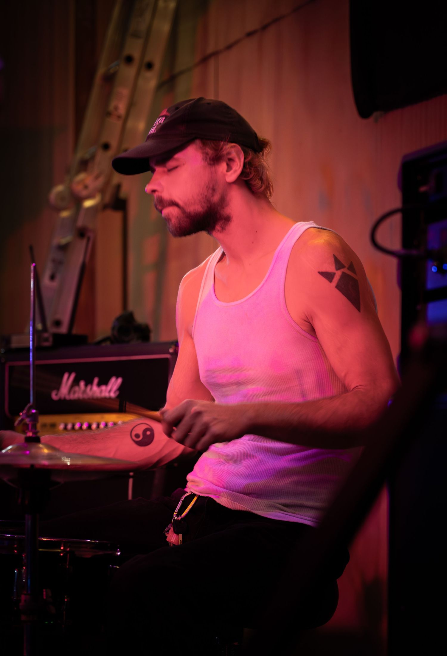 Musician in white tank top performs on stage under pink and purple lighting at a live music venue.