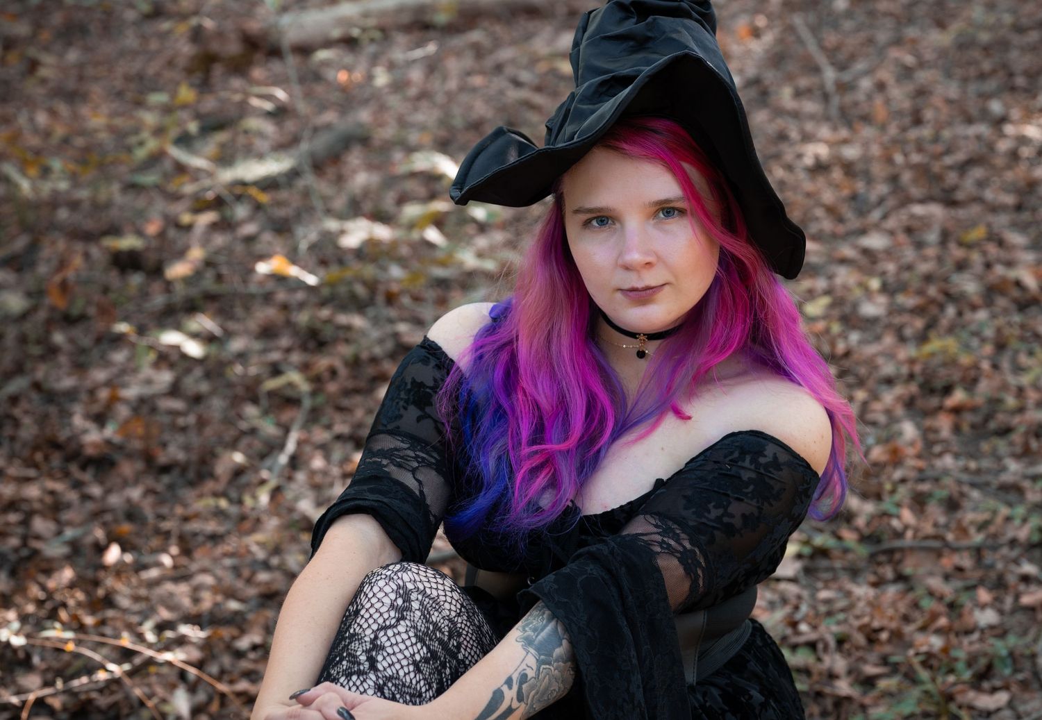 A person with vibrant purple hair wearing a black dress poses outdoors among fallen autumn leaves.