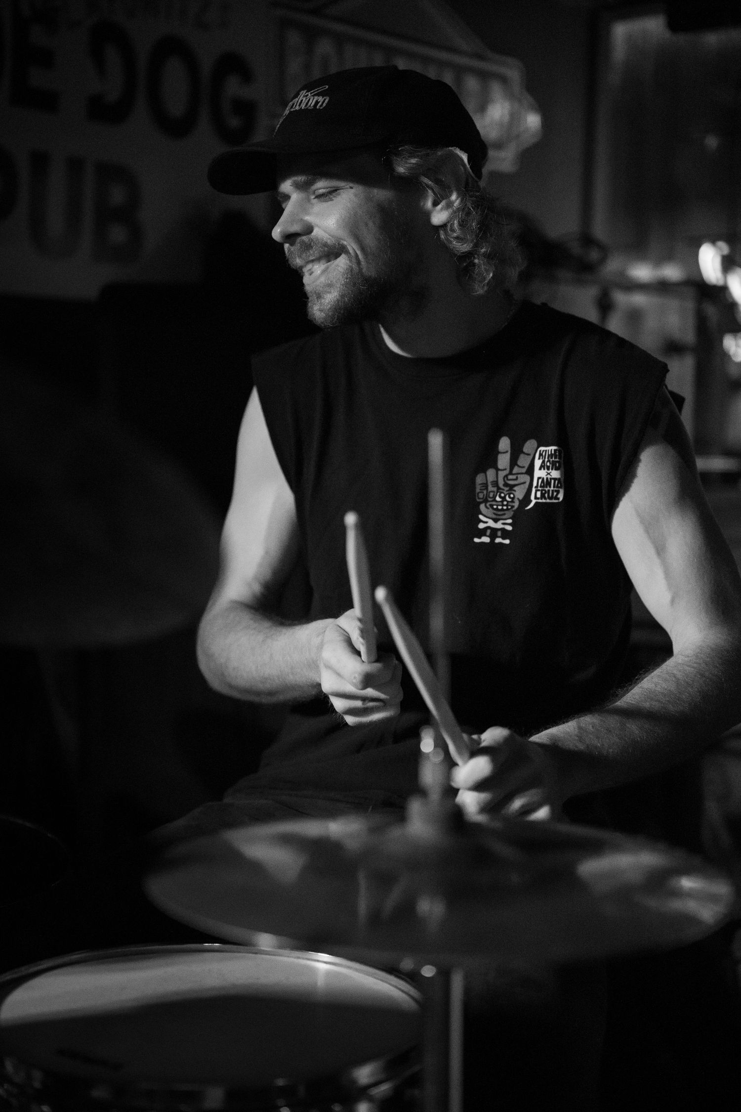 Black and white photo of a smiling male drummer playing drums at a live music venue.
