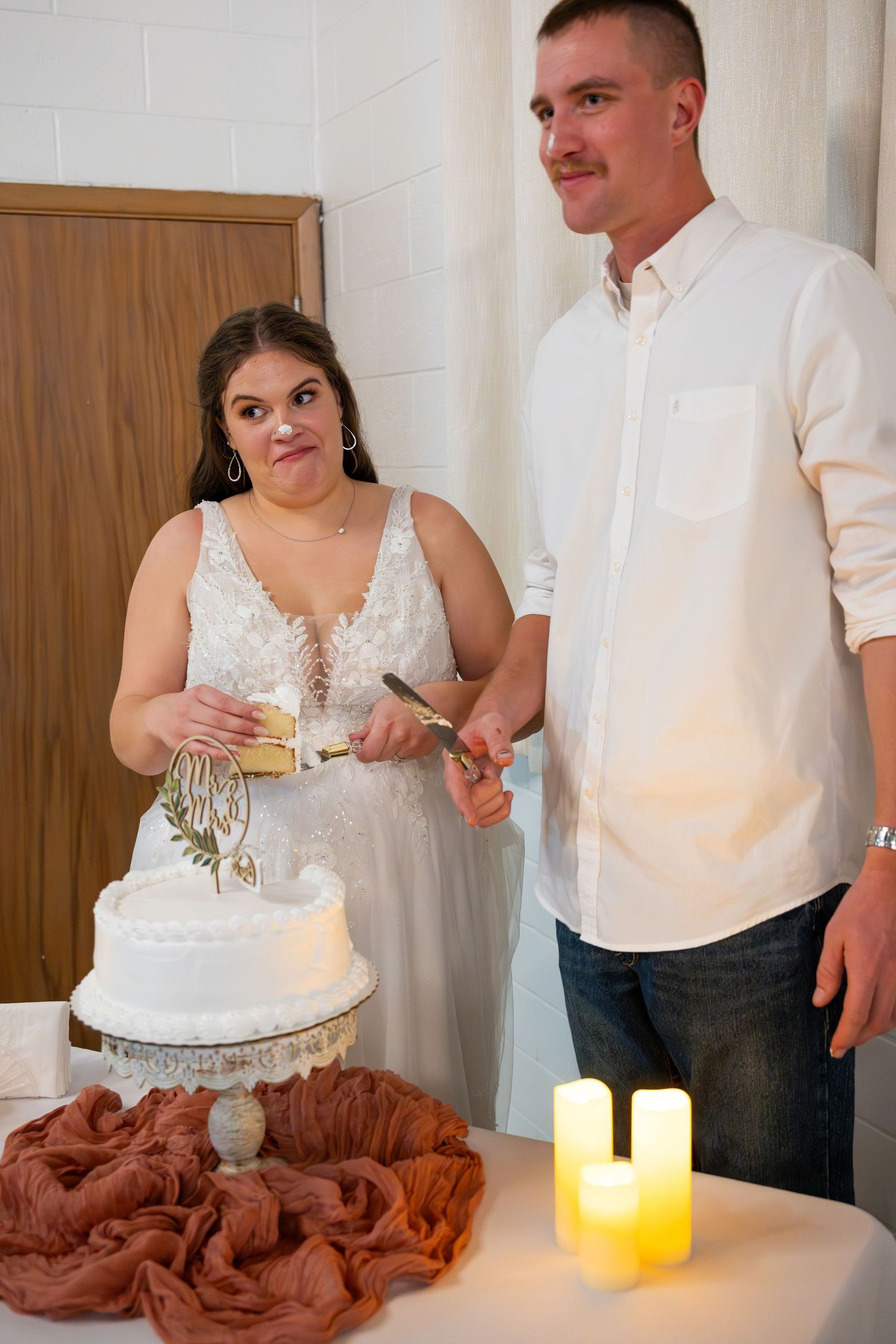 A couple cuts their white wedding cake together in an indoor setting with warm lighting.